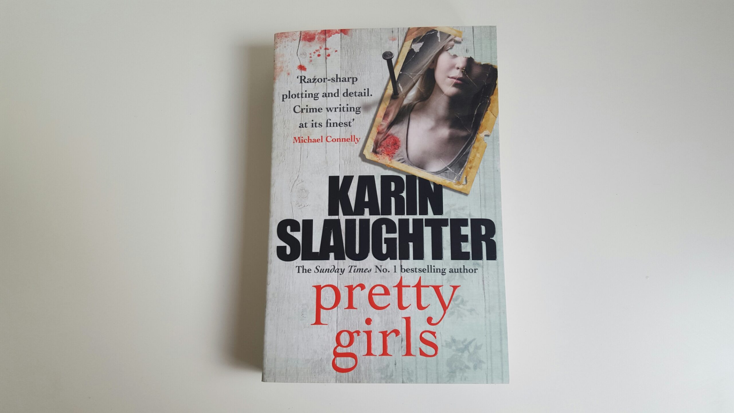 Pretty Girls by Karin Slaughter. The cover of the book appears to be white wooden wall that has a torn photograph of a woman nailed to it. By the photo appears to be blood splatter.
