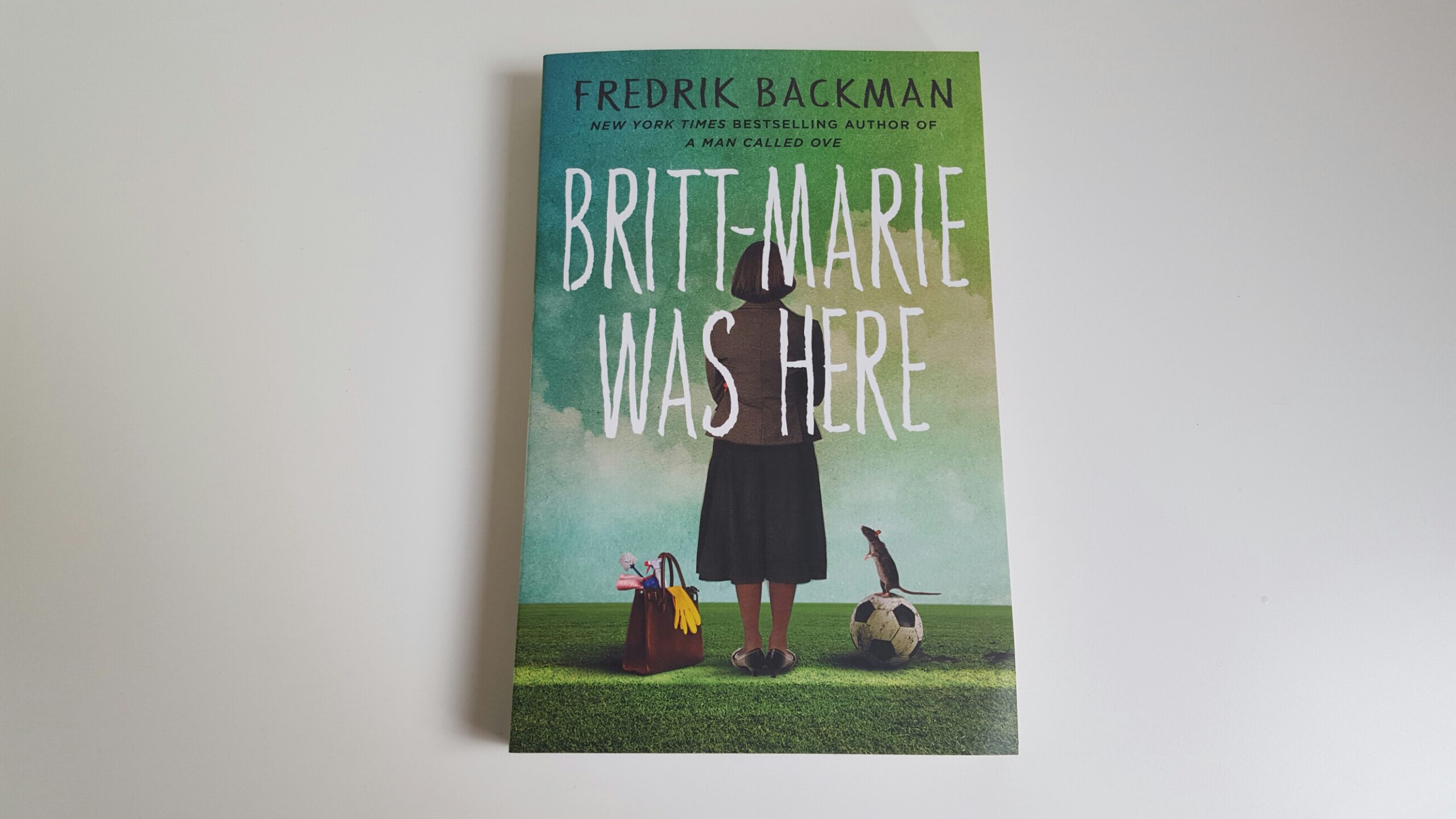 Britt-Marie was Here by Fredrik Backman. The cover shows a woman wearing a purple long-sleeve shirt and a black skirt with her back towards the reader. Next to her on the left is a brown bag with cleaning supplies sticking out of it. On her right is a soccer ball wit ha mouse standing on it.