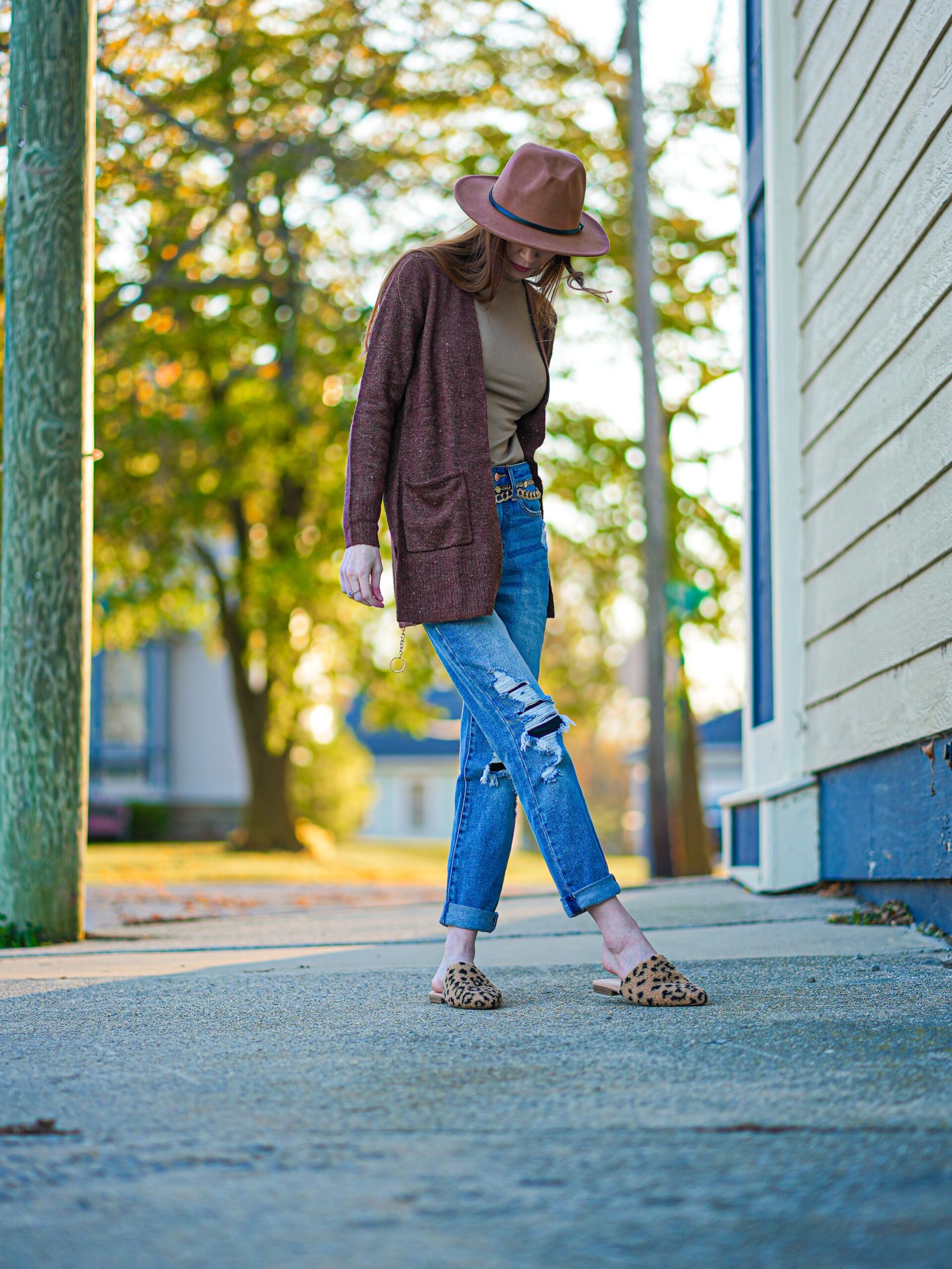 A woman wearing jeans with a light brown shirt, brown cardigan and a hat. 