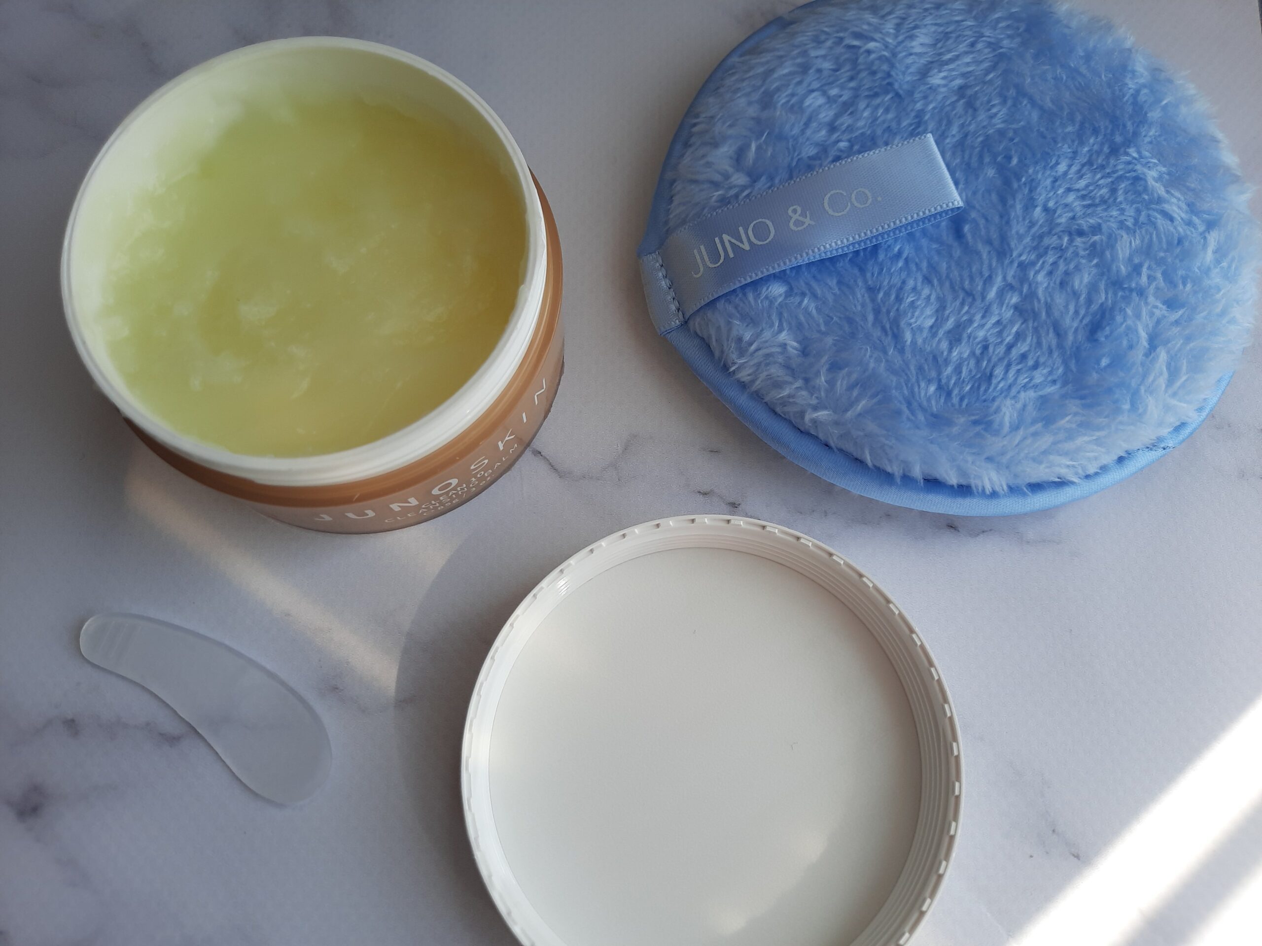 The JUNOCO Clean 10 Cleansing Balm opened to show off the balm as well as the spatula that is included. The cleansing cookie is next to the balm