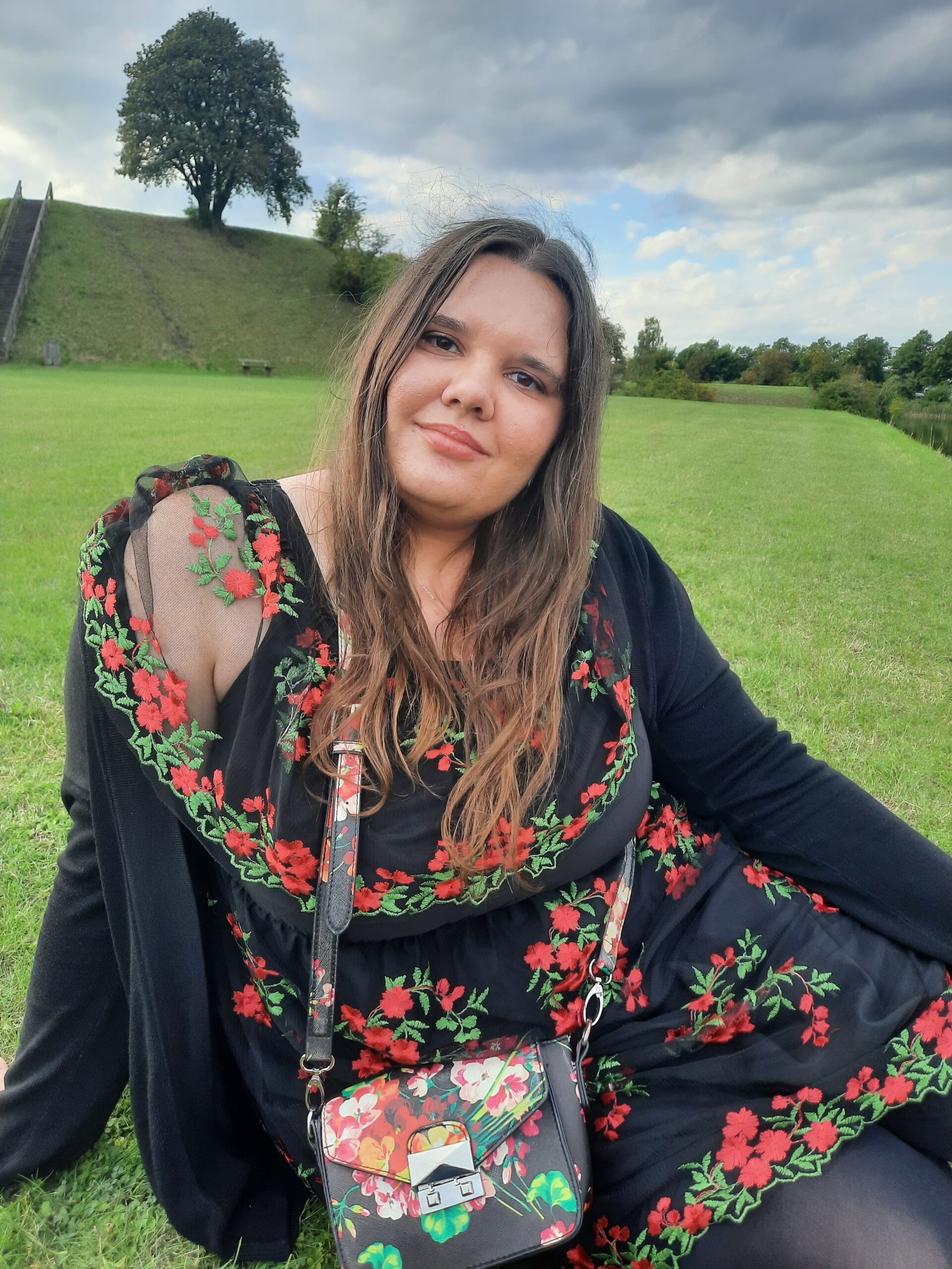 A photograph of myself wearing a black dress with red flowers and sitting on the grass and smiling