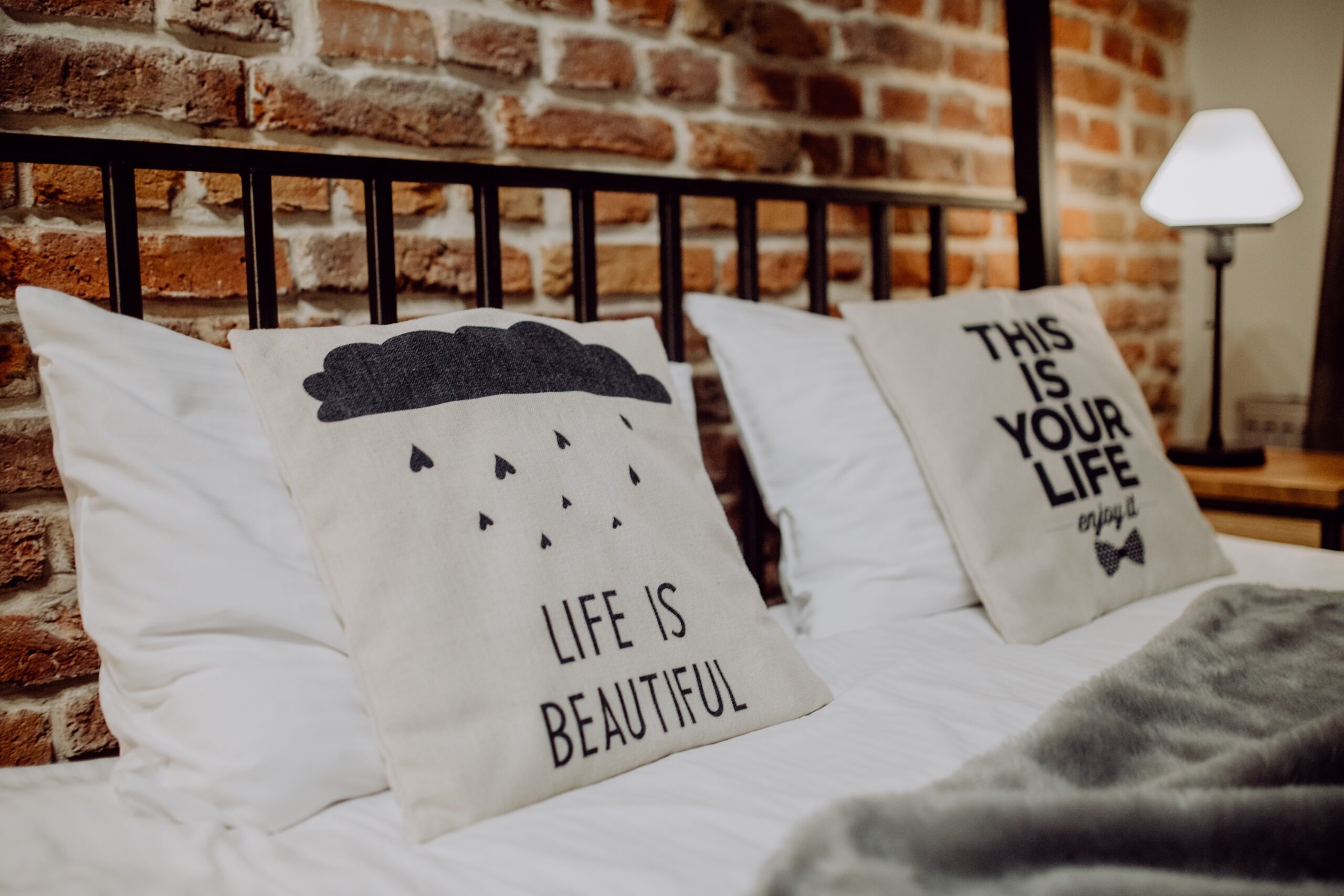 A bed with a black metal frame. On the bed, are two pillows that state life is beautiful and this is your life enjoy it. There is also a grey fluffy blanket laying on the bed.