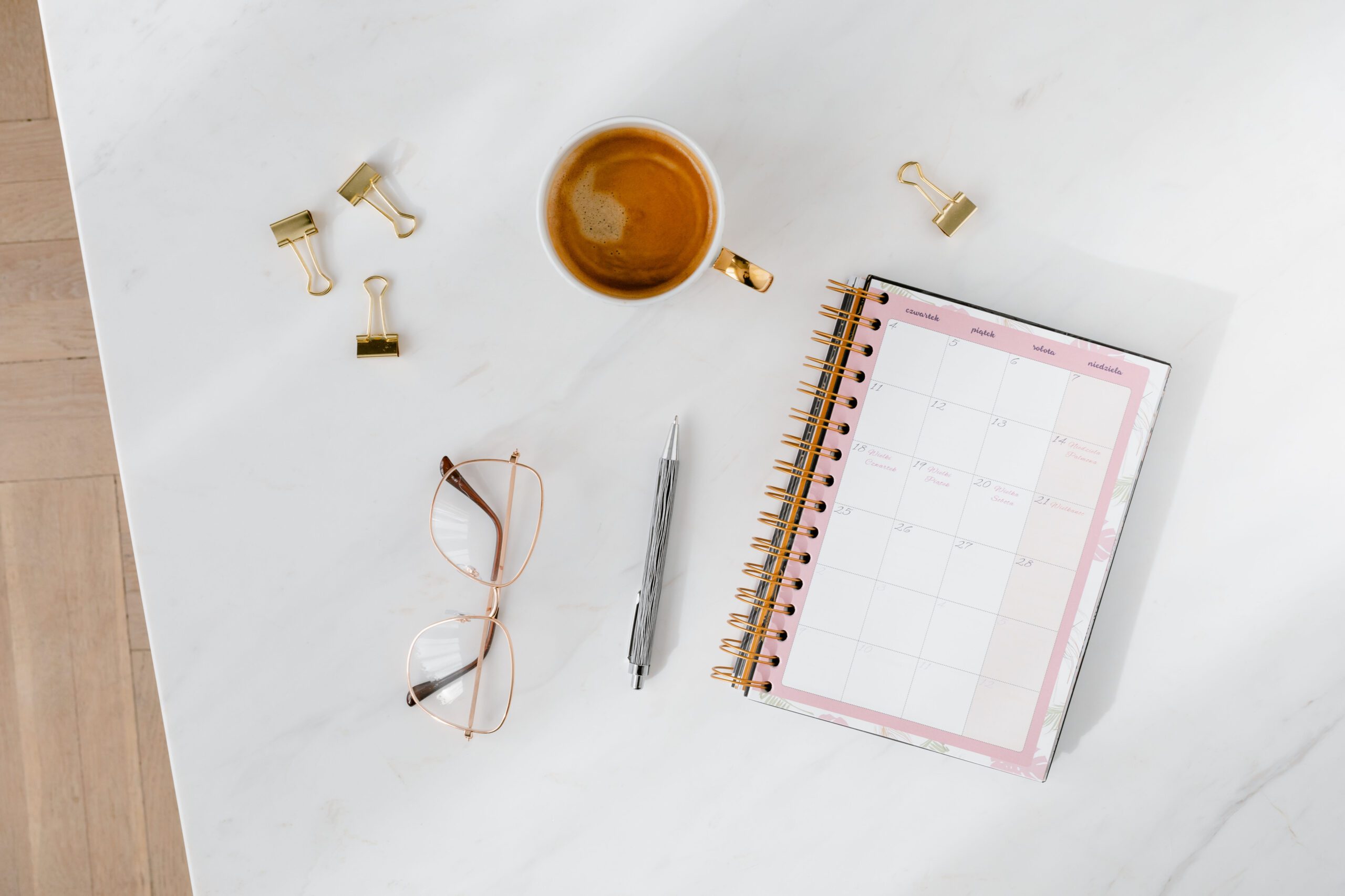 A spiral notebook laying on marble surface that is opened to a calendar page. Next to the notebook is a grey pen, a pair of glasses, and a cup of coffee.