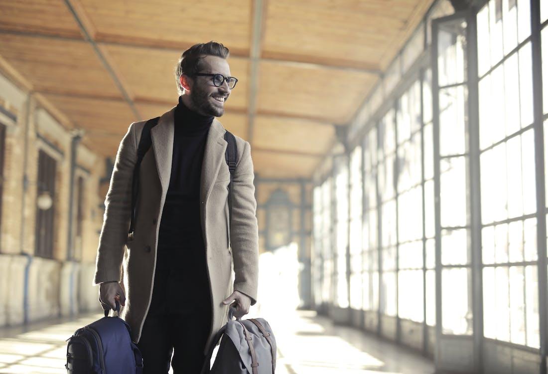 A man holding two pieces of blue and grey luggage while wearing a backpack and standing in what appears to be an airport. The man is wearing a black shirt and pants, a tan coat, and black glasses. The man is looking to the side and smiling.