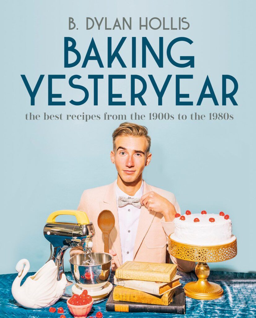 The cover of Baking Yesteryear by TikToker and YouTuber B. Dylan Hollis. The book shows Dylan wearing a pink suit jacket and white buttoned-up top with a bowtie. Dylan has blond hair and is holding a wooden spoon. On the table is a white cake on a gold cakestand, a stack of four old books, a small stand mixer, a white swan statue, and a bowl of cherries. 