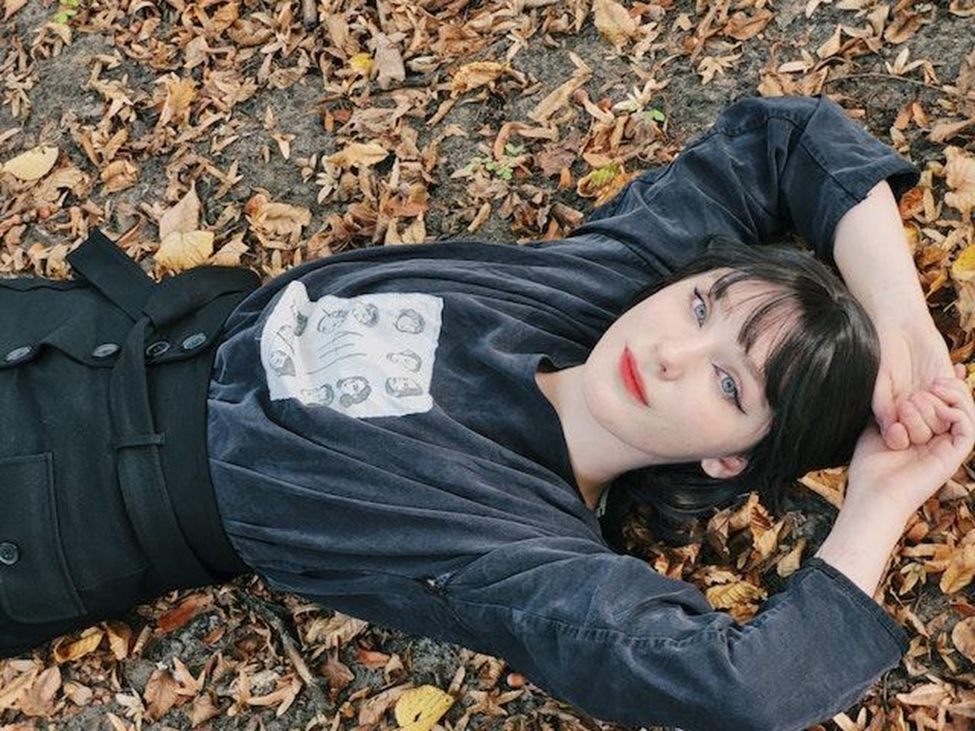 A woman lying on the ground with fallen leaves around her. She is wearing black eyeliner and red lipstick and wearing a black longsleeve shirt and black bottoms. 