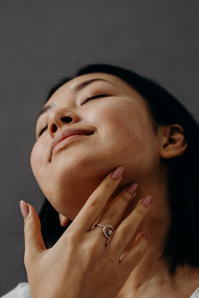 A headshot of a woman with her eyes closed and touching her face. The woman has short black hair and her nails are painted a dusty pink color. She has is wearing a moon and star silver ring on her middle finger. 