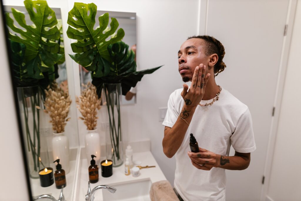 A man applying skincare to his face in the bathroom. The man has brown hair and is wearing a white t-shirt and gold necklace. He also has small tattoos on his arms. 