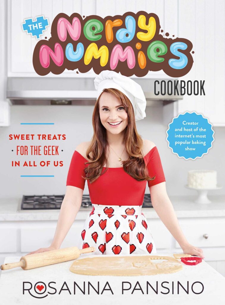 The cover of The Nerdy Nummies Cookbook by Rosanna Pansino. The cover shows Rosanna standing in a white kitchen. She has brown hair and is wearing a white chef hat, red top, and white skirt with red pixel hearts. In front of her is rolled-out cookie dough and a cookie cutter. 