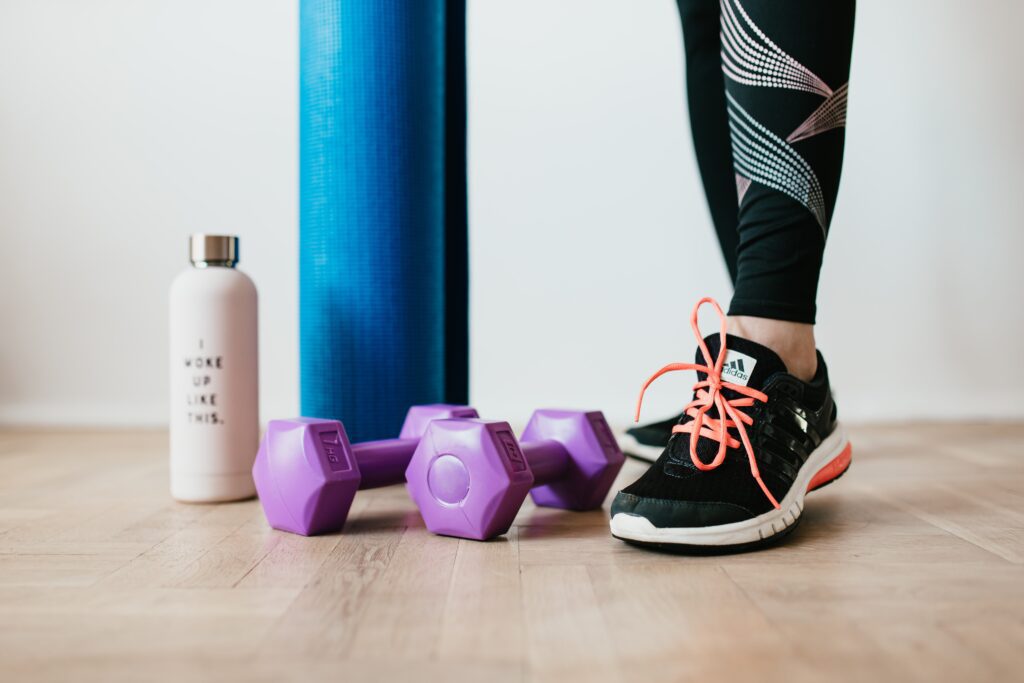 A woman wearing black leggings and black sneakers standing next to purple dumbells, a blue rolled up yoga mat and a white water bottle.