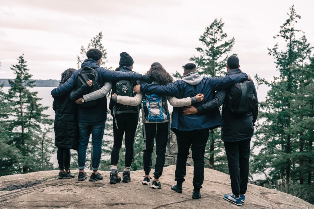 A group of 6 people holding each other with their backs faced towards the camera and looking out on the edge of a cliff. 