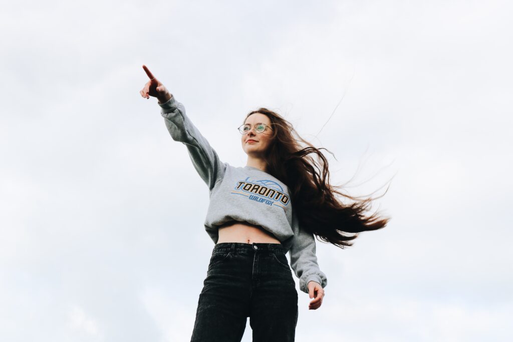 A woman with long brown hair wearing a grey Toronto Wildfox sweater and black jenas pointing with her right hand