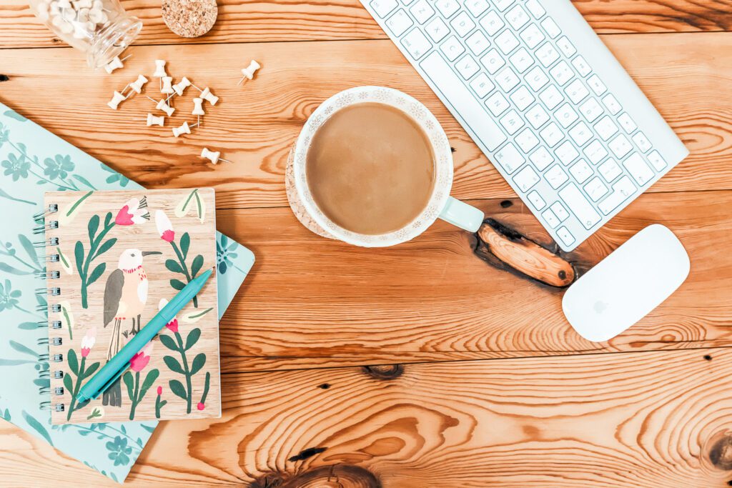 A white keyboard and mouse, a cup of coffee, and a notebook with a bird and floral pattern with blue pen laid on top of a wooden desk.