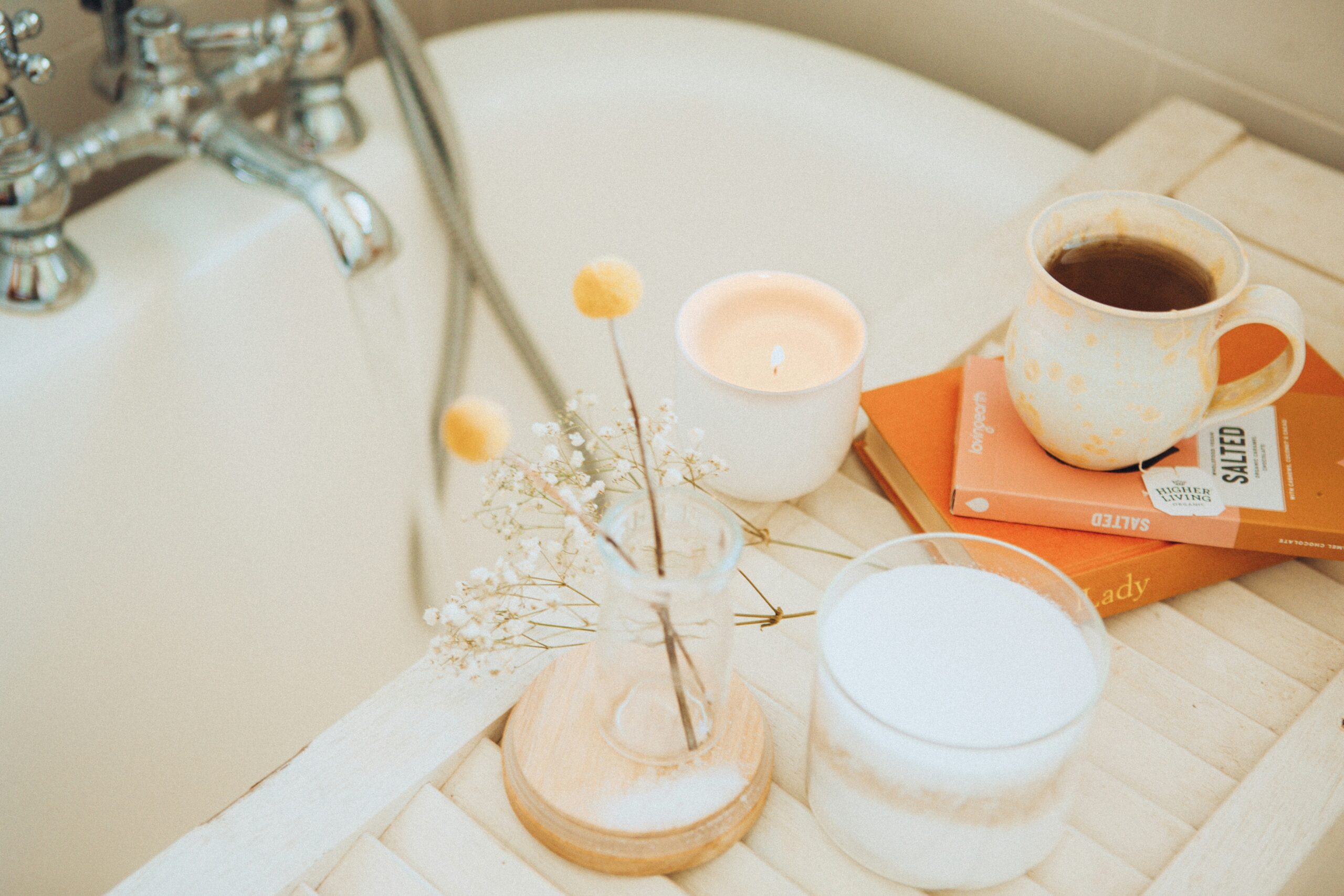 A bath filling with water with a tray that has a lit candle, a mug filled with tea, and books.