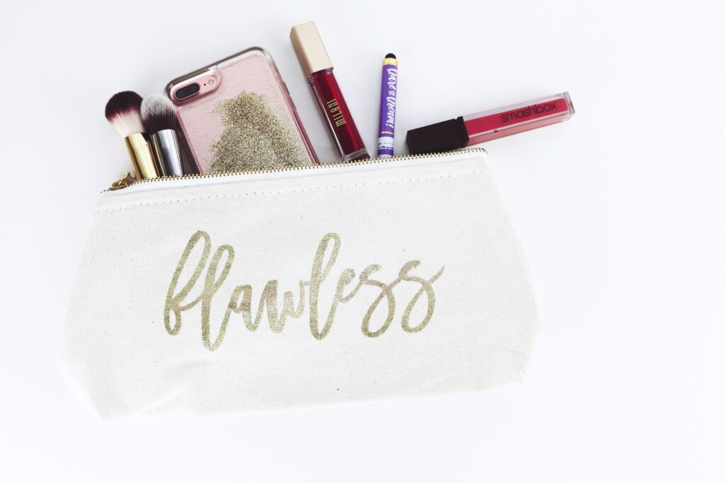 A canvas makeup bag with 'flawless' written in cursive gold letters. In the makeup bag are liquid lipsticks, makeup brushes, and a phone.