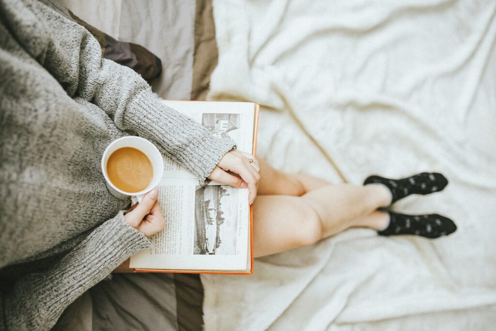 A woman wearing a grey sweater and reading a book while drinking a cup of coffee