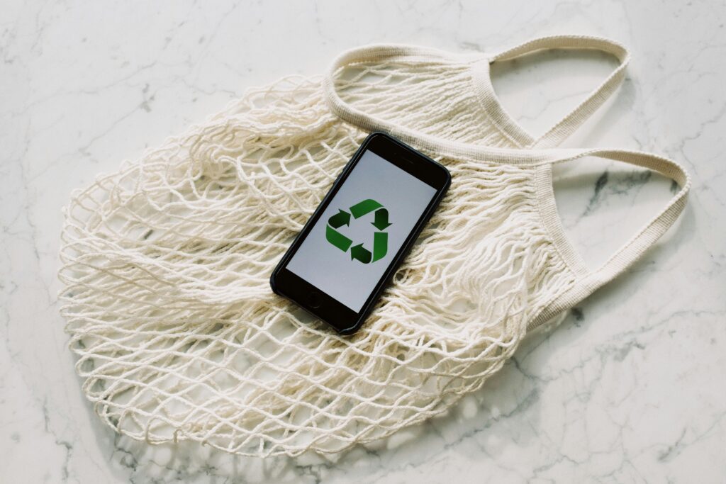 A cellphone with the green recycling symbol laid on top of a mesh bag.