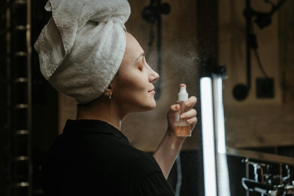 A woman wearing a black shirt with with her hair wrapped in a white towel and spraying a facial mist on her face