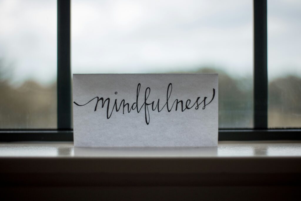 A small white sign that states mindfulness in a black curvy font. The sign is in front of a window on a small shelf.