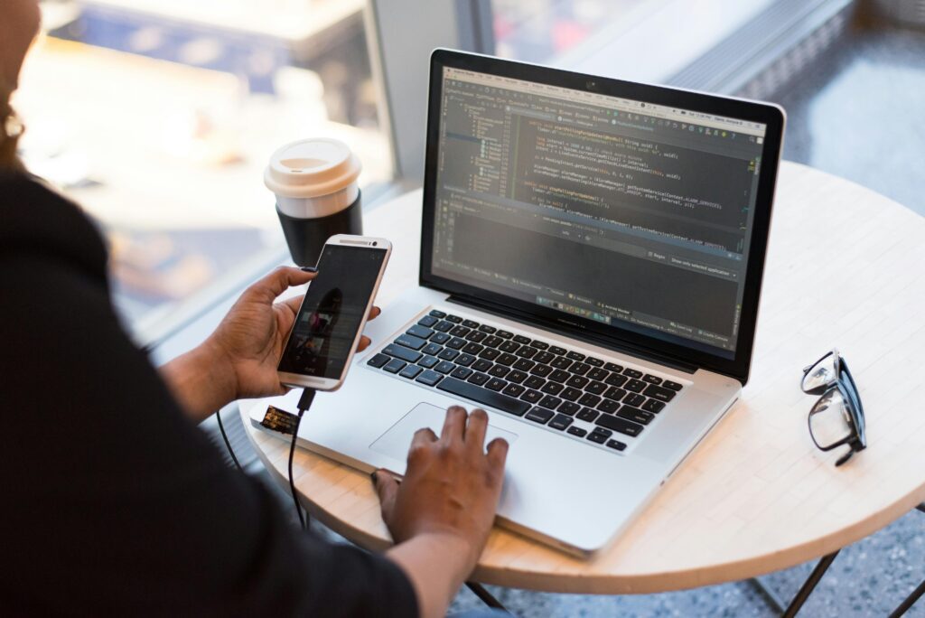 A person holding a smartphone that is connected to their laptop and working on coding