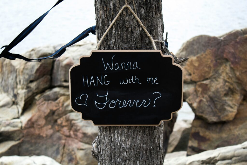 A small chalkboard sign hanging on a tree that has "Wanna hang with me forever?" written on it