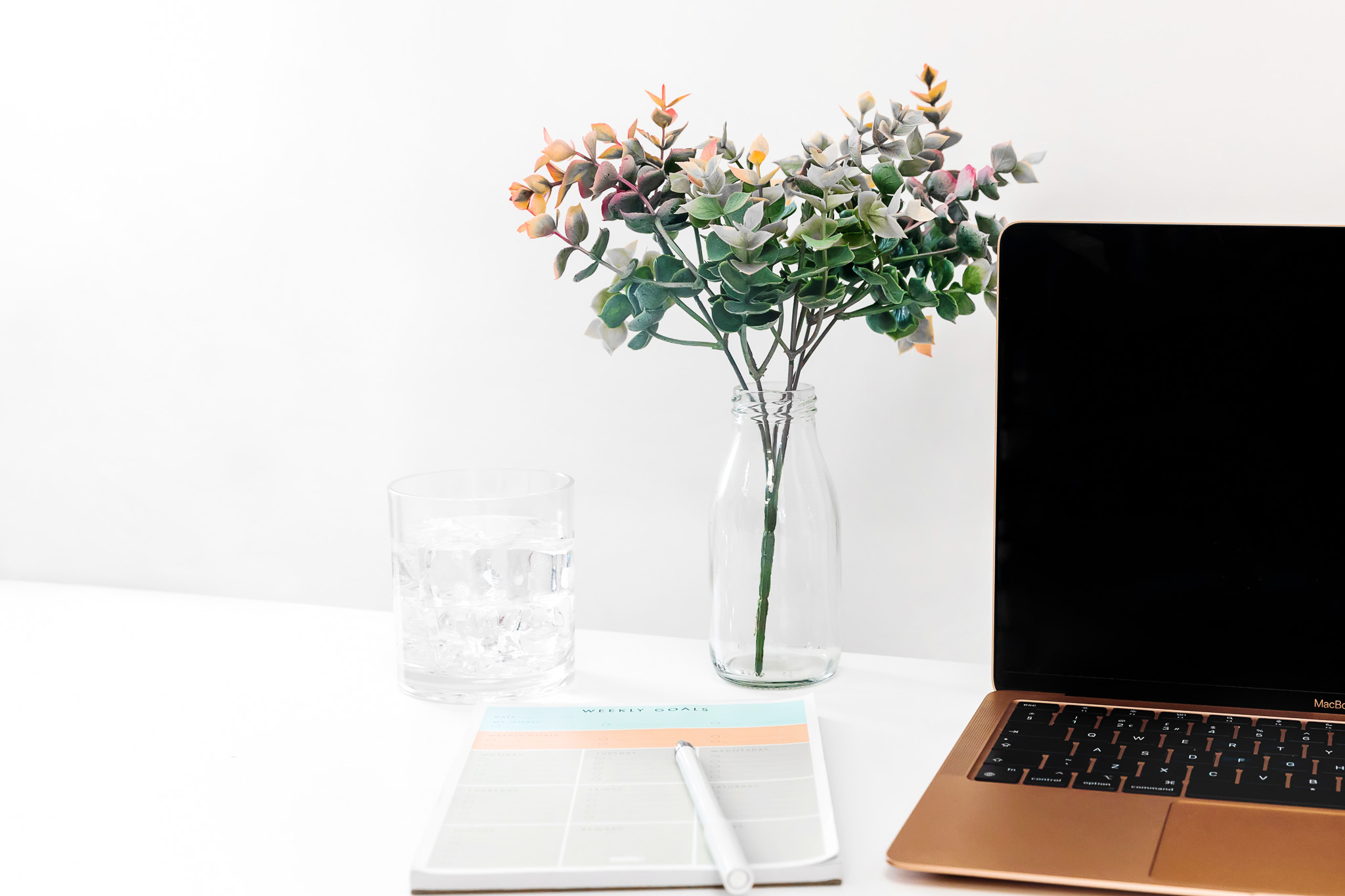 A weekly goal list on a white table next to a gold laptop and glass vase with plants