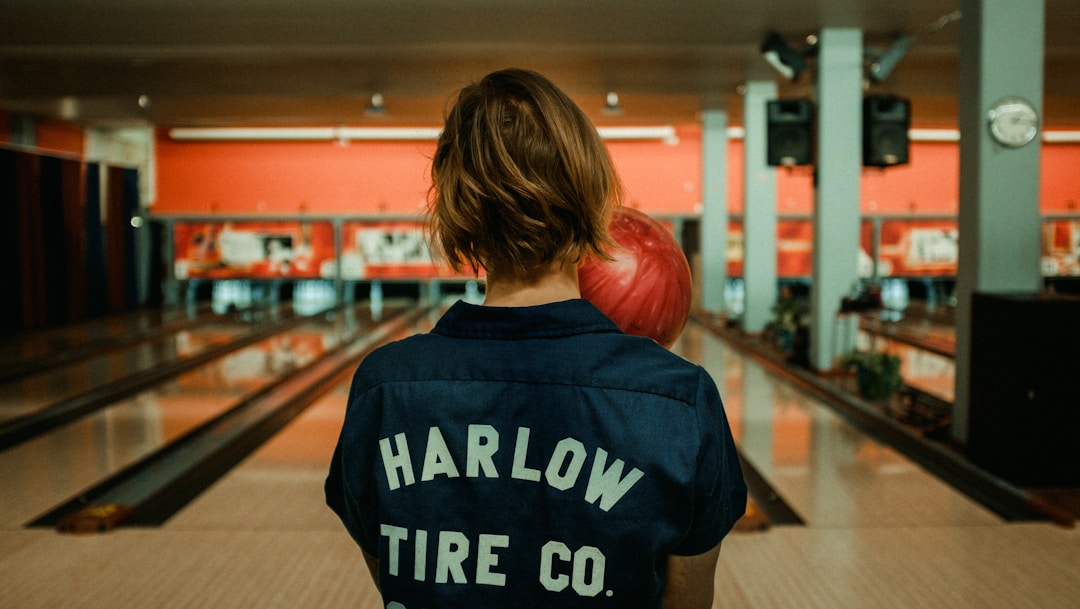 A young woman wearing a blue bowling shirt with the words "Harlow Tire Co." on the back and holding a red bowling ball in front of a bowling lane
