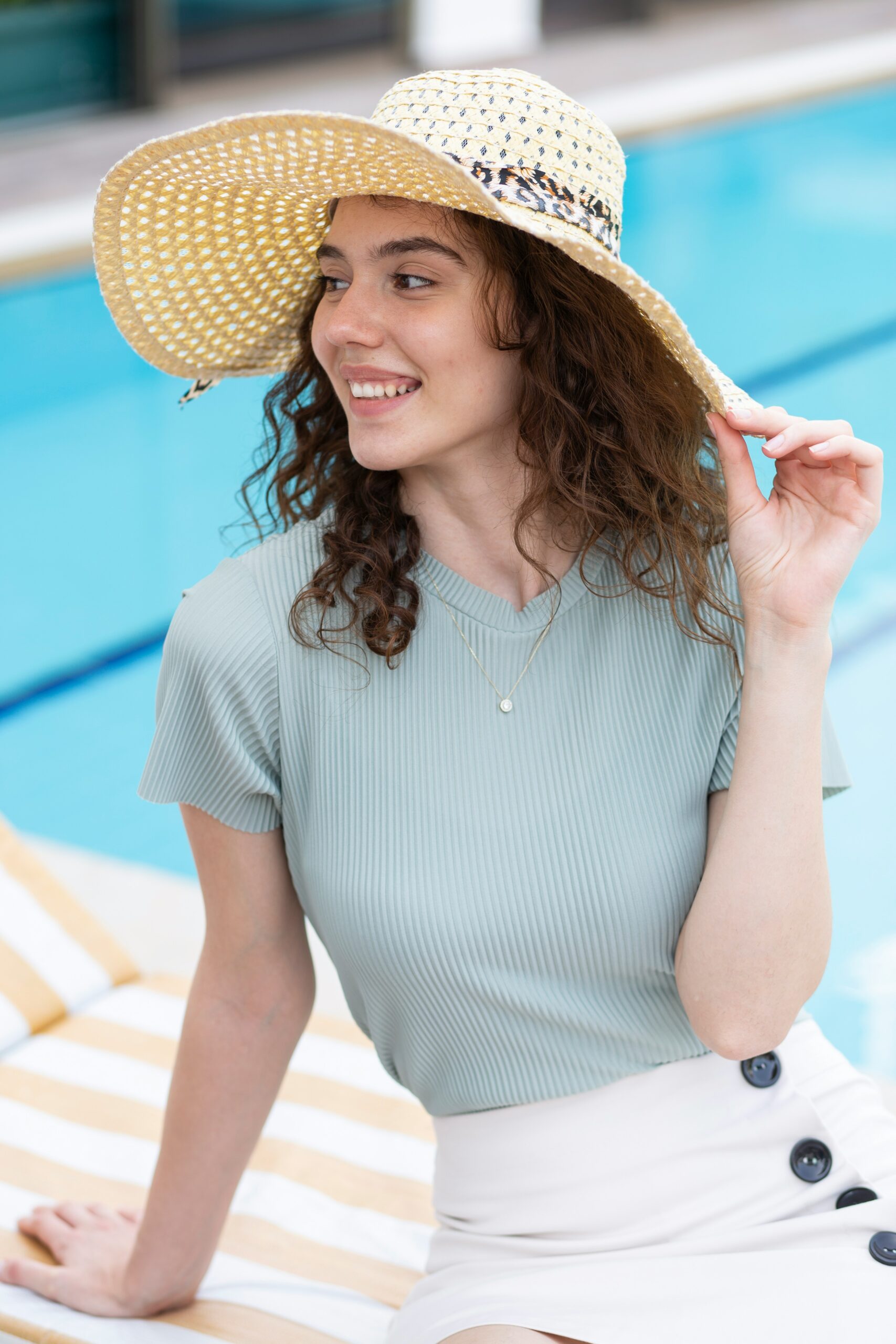 A woman with brown curly hair sitting beside a pool and wearing a creme sunhat. a light blue short-sleeved shirt. and a white skirt with black buttons.