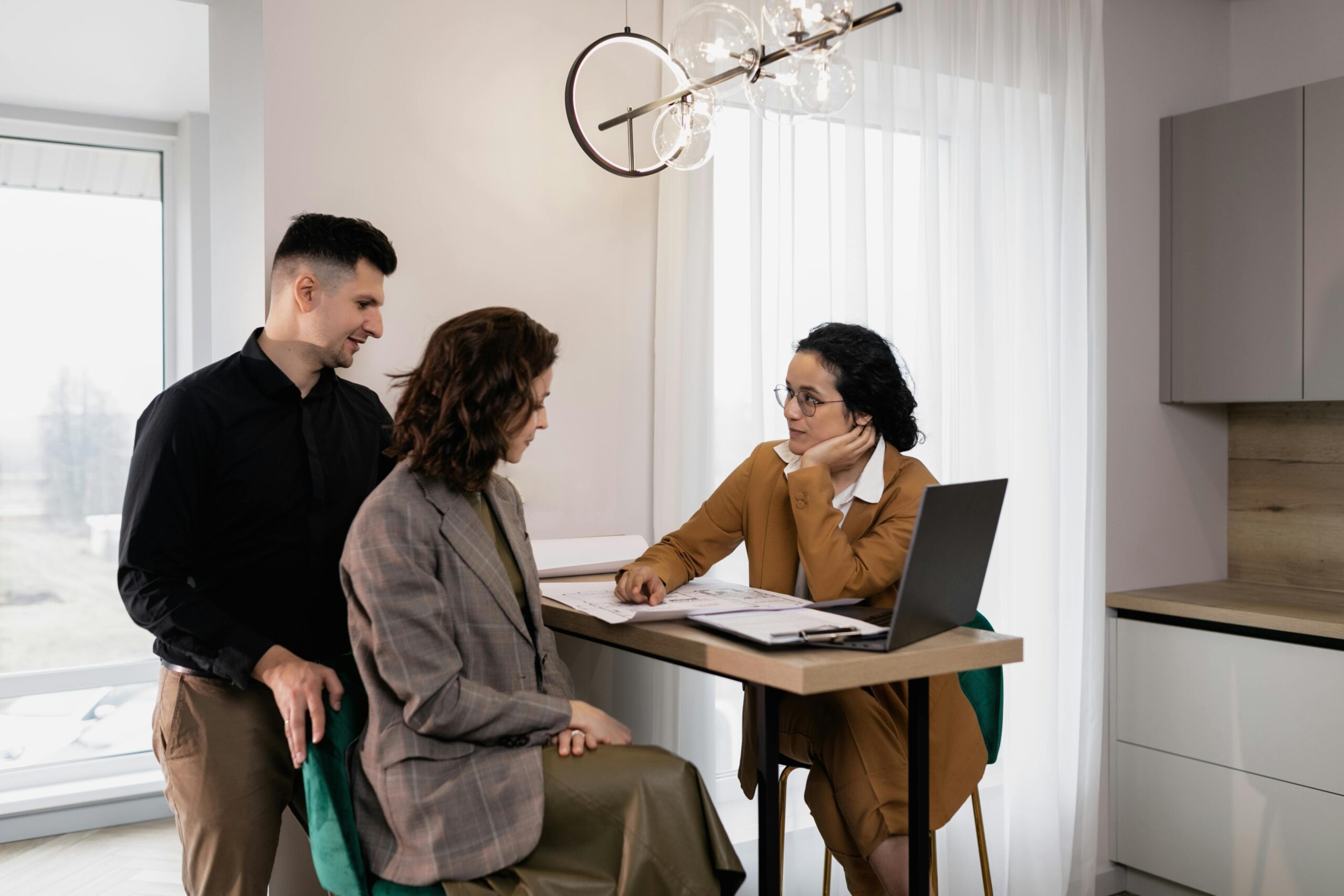 A man with short black hair wearing a black shirt and brown pants standing behind a woman who is sitting a chair. The woman has curly brown hair and is wearing a light brown tweed jacket and a brown skirt. They are meeting with a real estate agent who has black hair and wearing a pant suit.