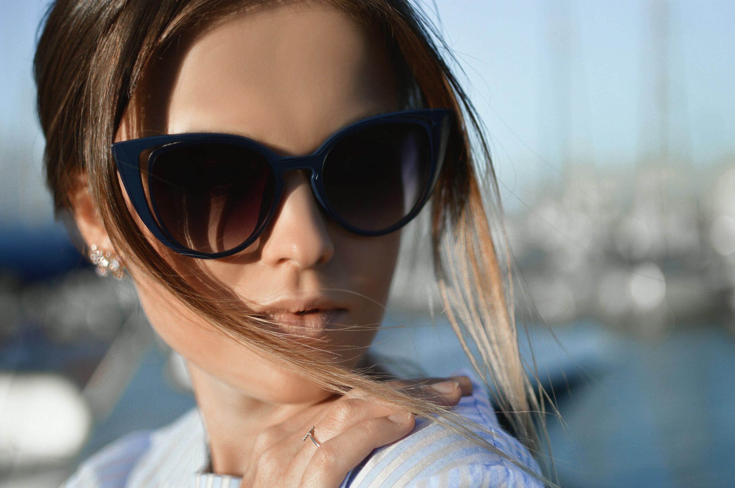 A close-up of a woman with brown hair wearing a black pair of sunglasses. Her hair is tied up and her hand is on her shoulder.