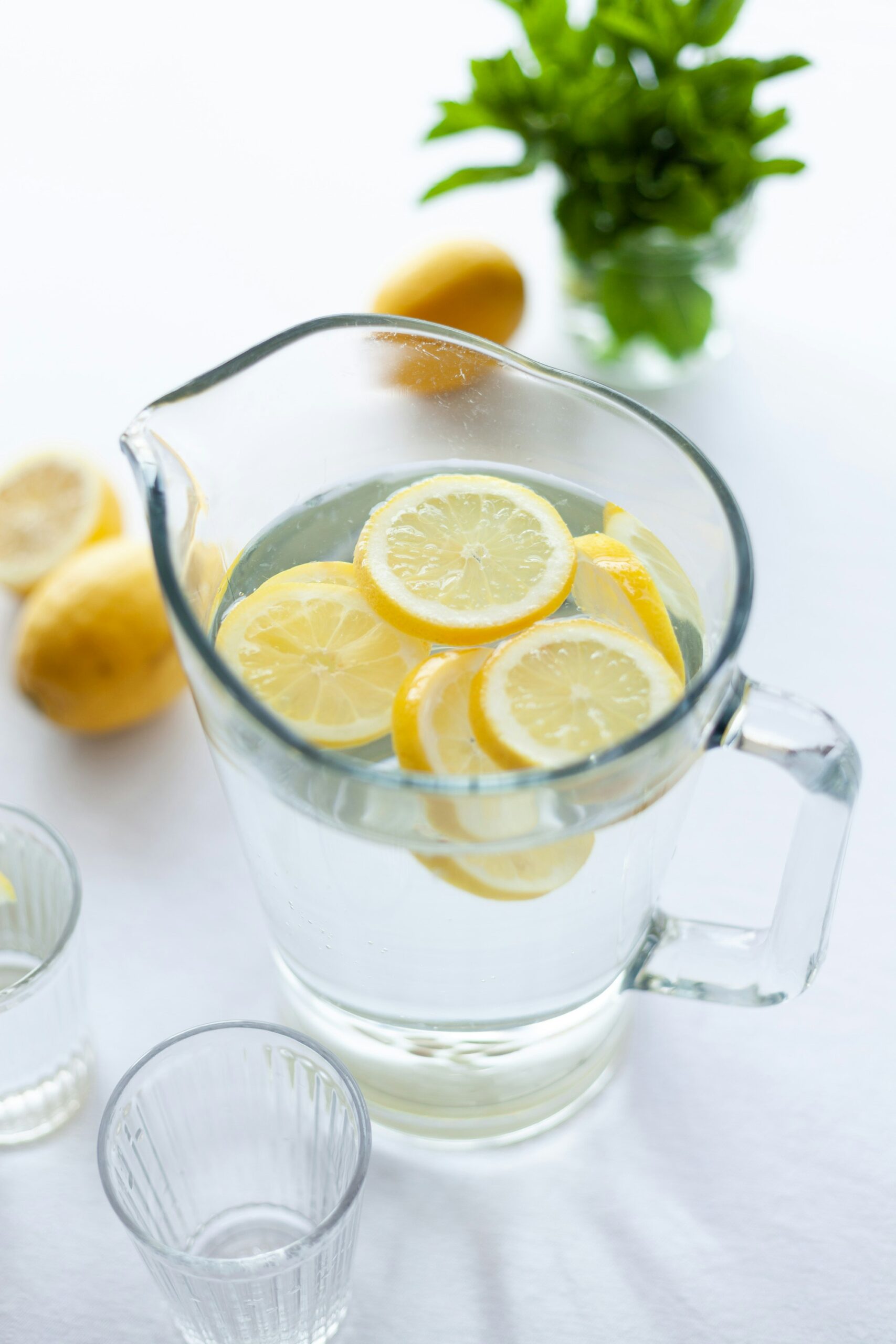 A glass pitcher filled with water and topped with lemon slices. Next to the pitcher are two glasses.