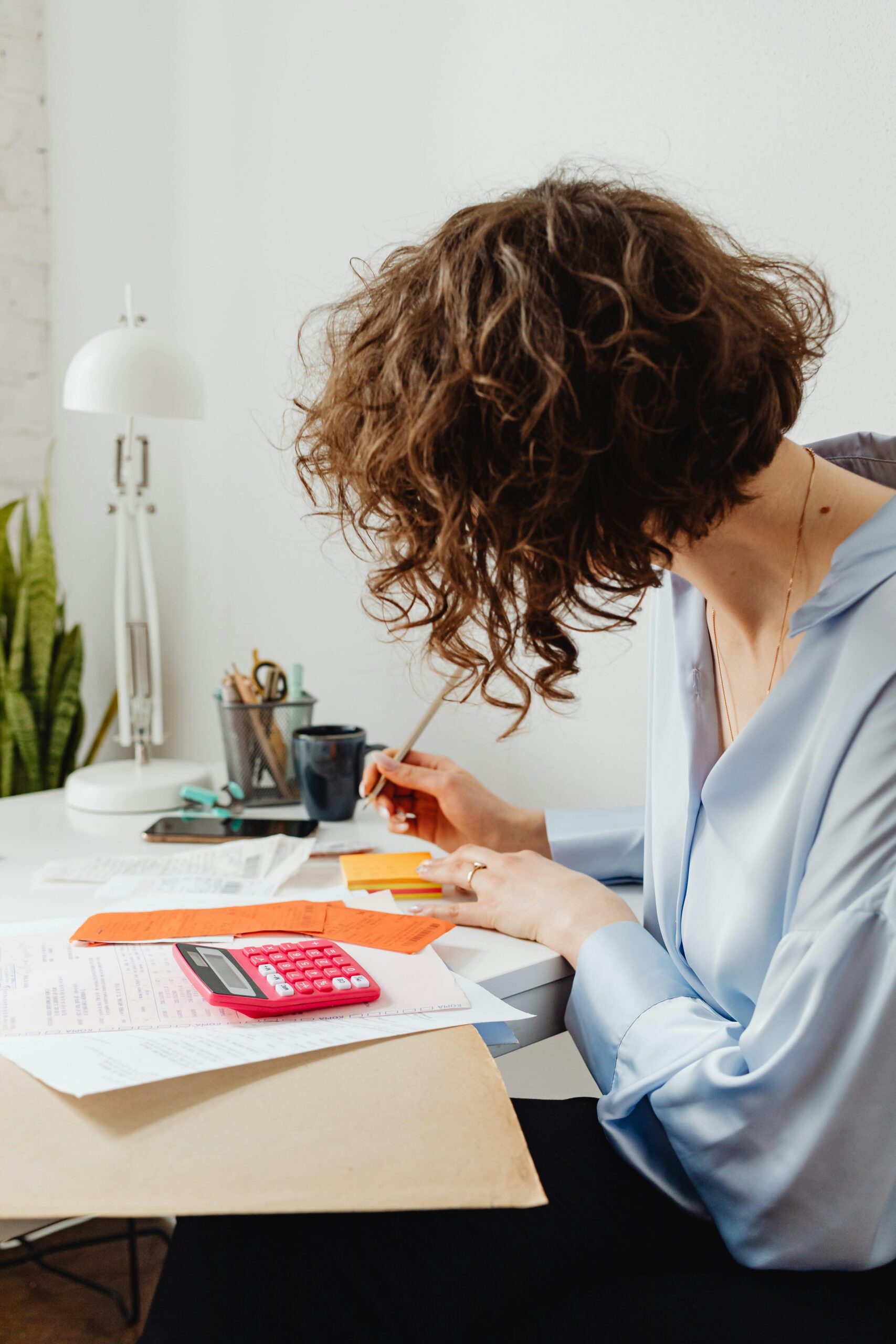 A woman with short wavy brown hair and wearing a light blue blouse sitting at a desk and working on a budget plan. On the desk is a red calculator, notepad, and various papers.