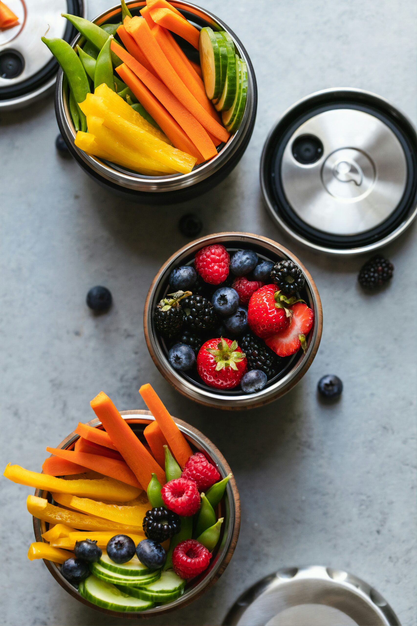 Snack containers filled with vegetables and berries. Two containers have carrot and yellow pepper sticks, cucumber slices, and sugar peas. The third container has strawberries, blueberries, blackberries, and raspberries.