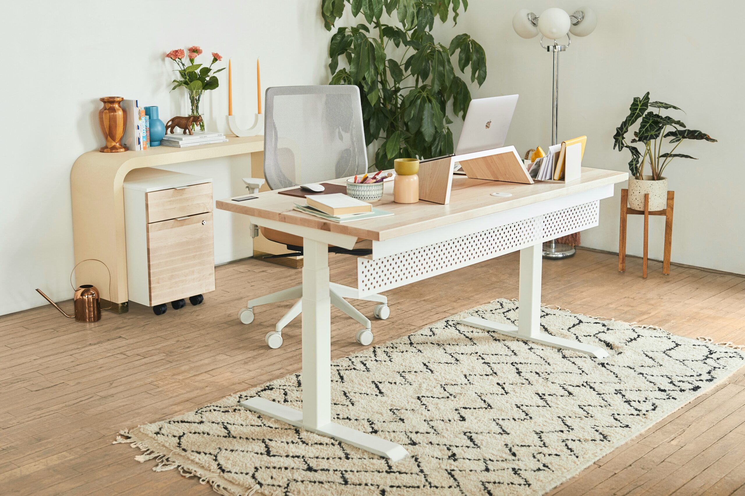 A home office with a laptop sitting on a white adjustable desk with a light wood top. In the office is an ergonomic rolling office chair and potted plants.