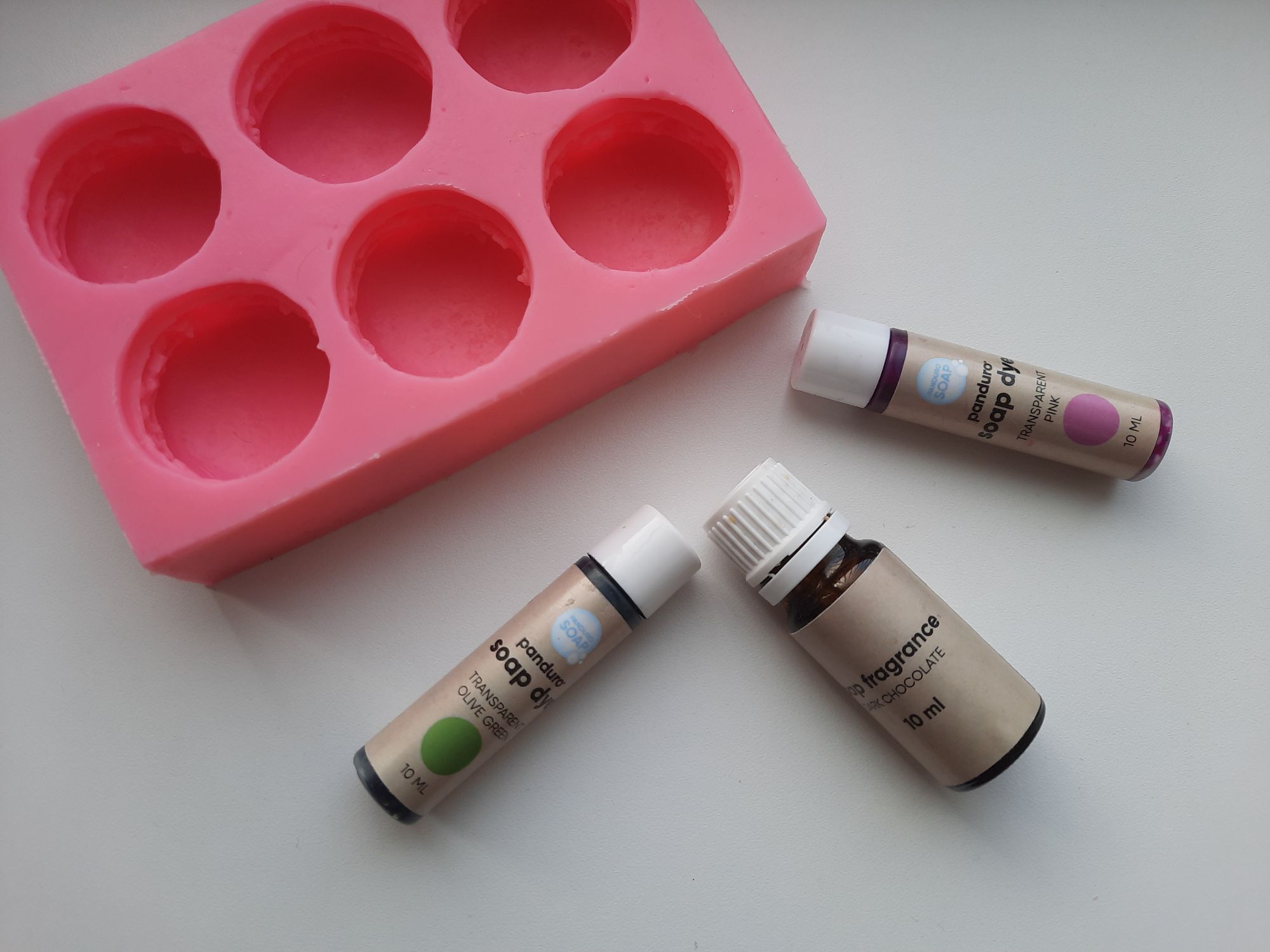 Some melt and pour soap making supplies including a silicone macaron soap mold, green and pink soap dyes, and a chocolate soap fragrance