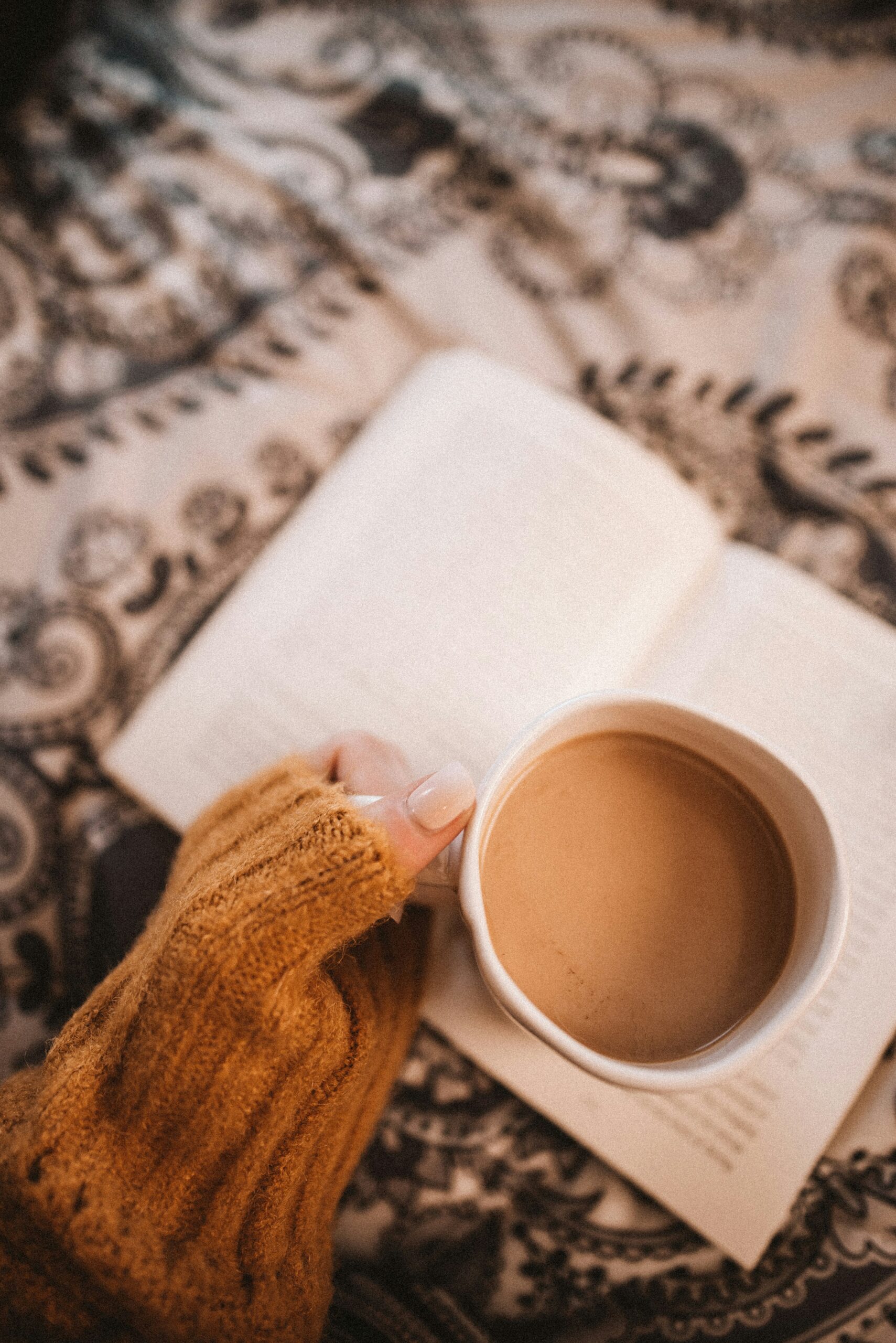 A close up of an orange sweater sleeve and a hand holding a coffee cup. In the background is an open book