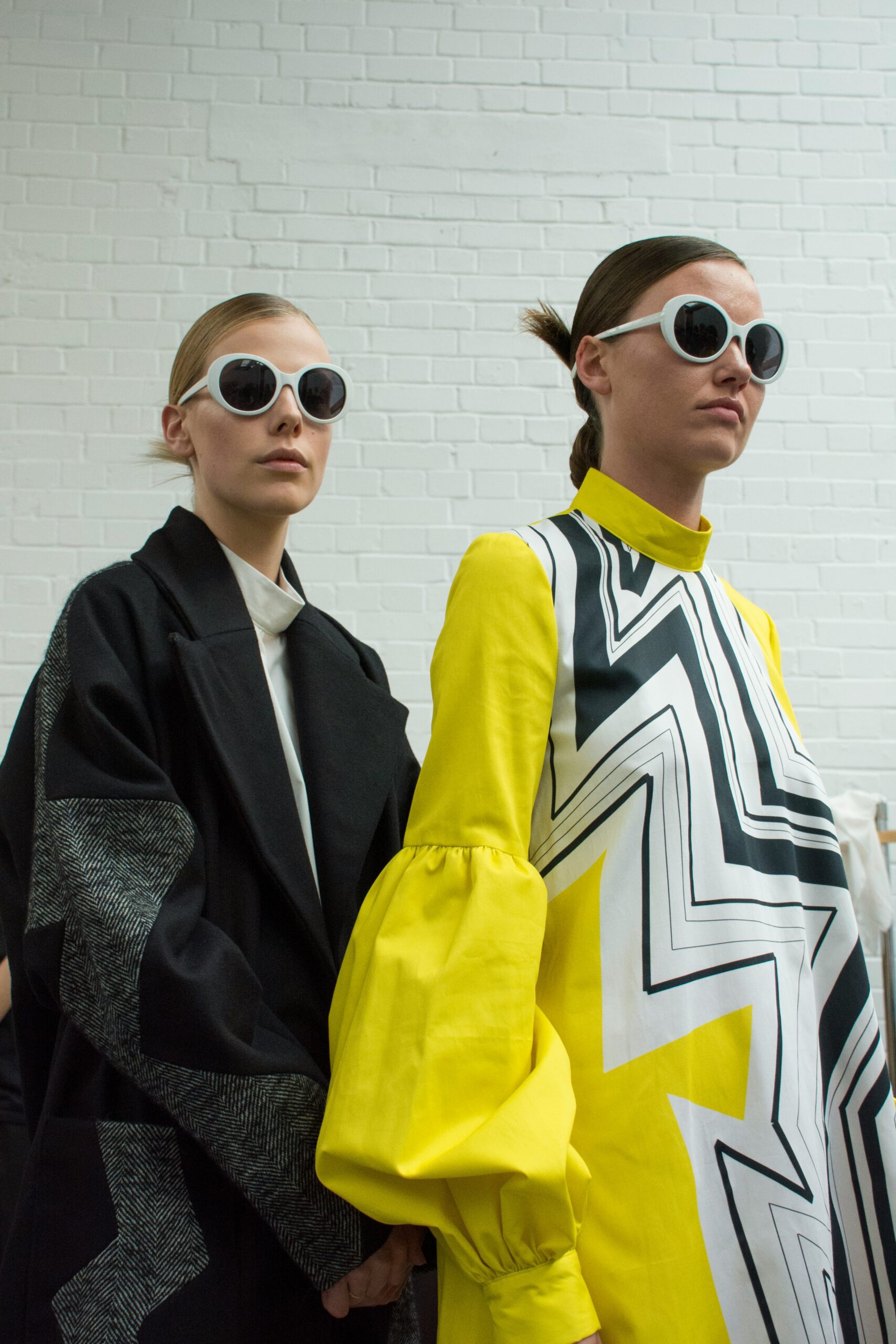 Two woman wearing maximalist clothing. One woman is wearing a yellow dress with a black and white zig zag pattern in the middle. The other woman is wearing an oversized black and grey jacket with a white undershirt. Both women are wearing round white sunglasses.