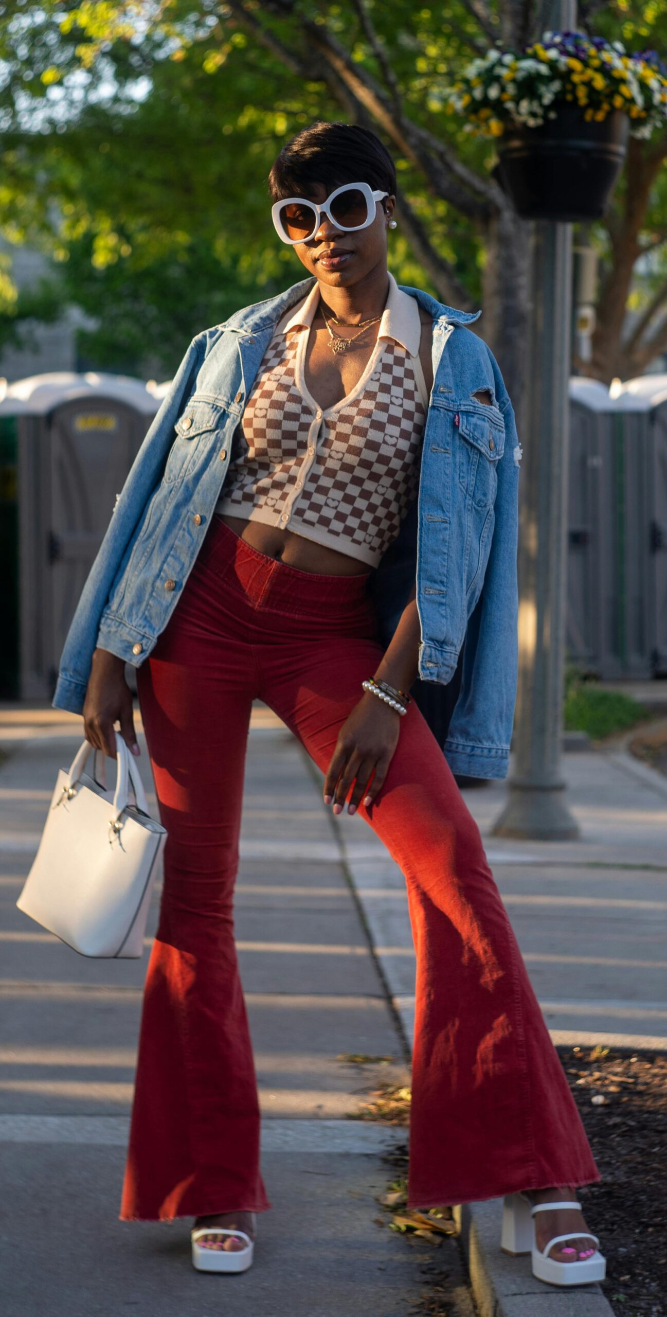 A woman wearing red flared pants with a white and brown checkboard pattern crop top. She is also wearing a denim jacket over her shoulders, round white sunglasses and white chunky heels.