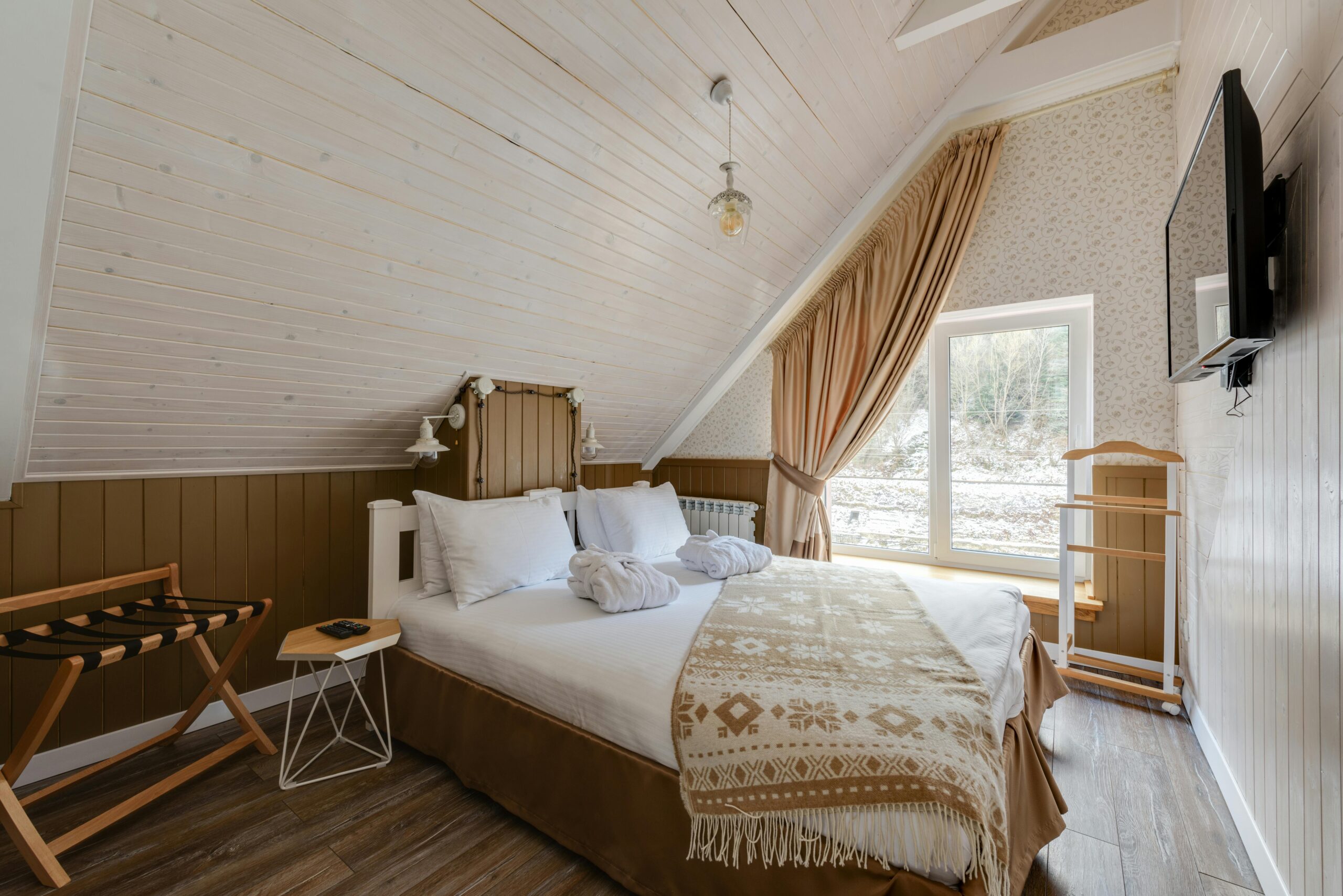 A bedroom with a bed with white sheets and a tan patterned throw blanket. Next to the bed is a small bedside table and luggage rack.
