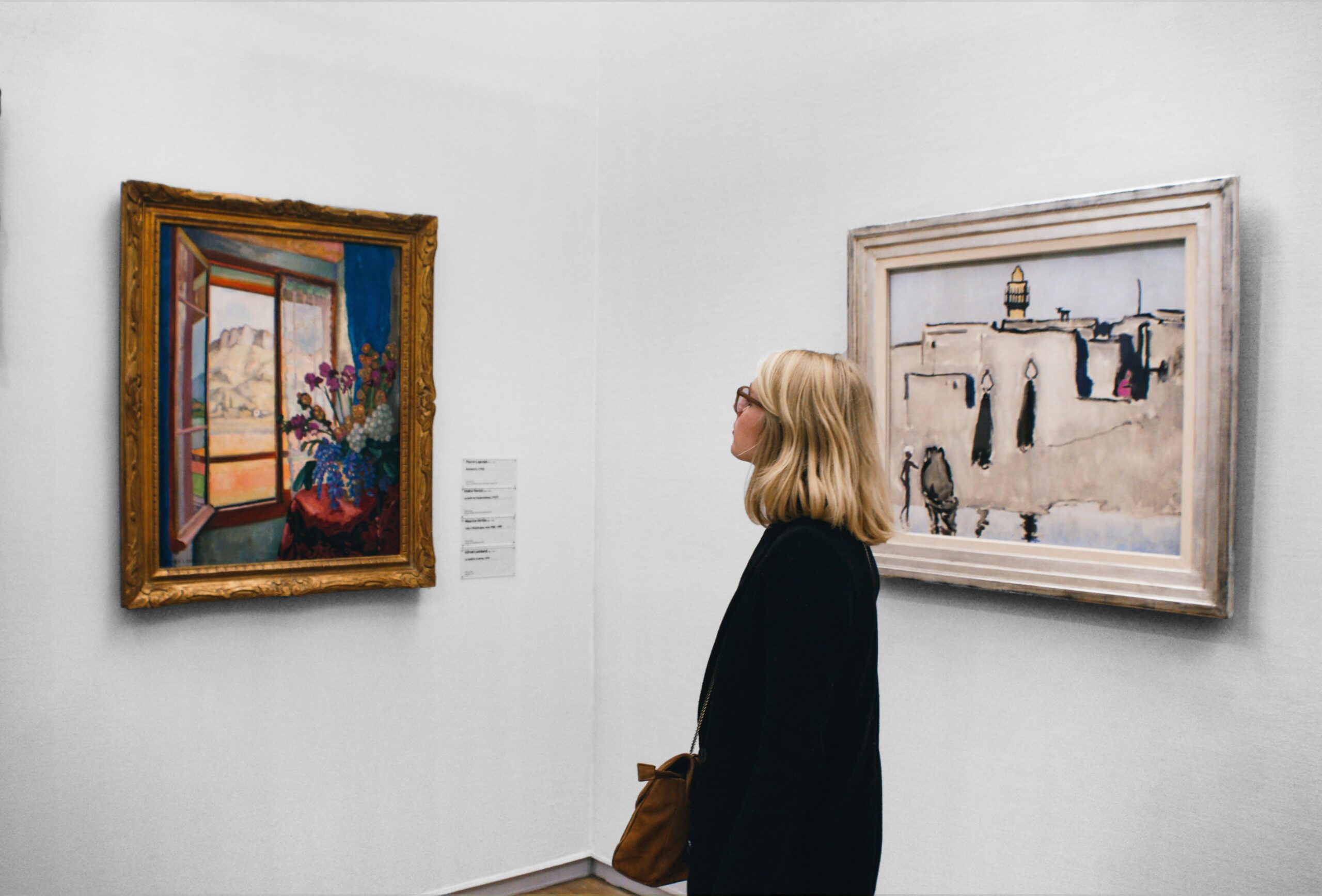 A woman with short blonde hair wearing glasses and a black jacket viewing a painting at a museum