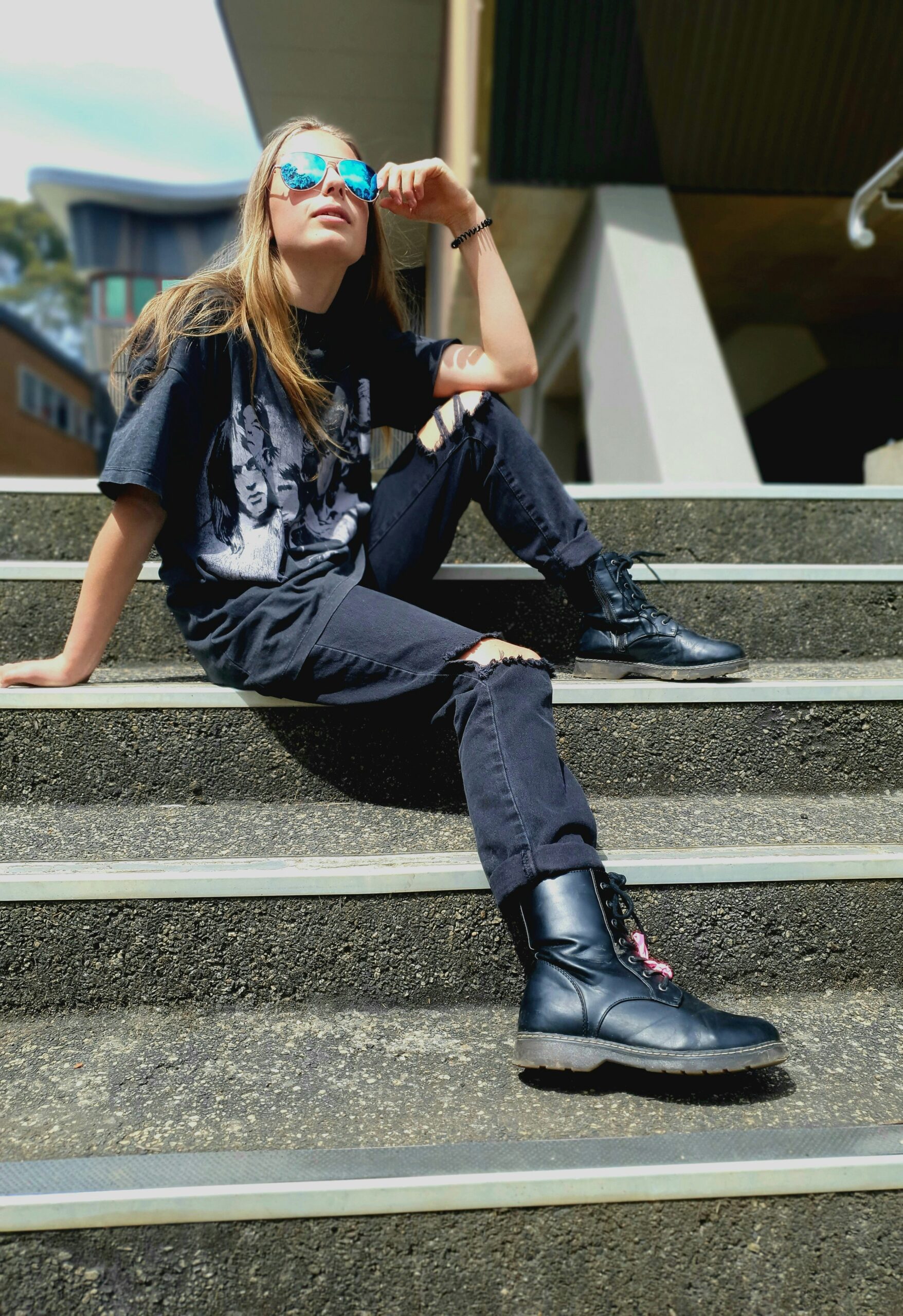 A young woman with blond hair wearing black ripped jeans, a black print tea, and sunglasses.