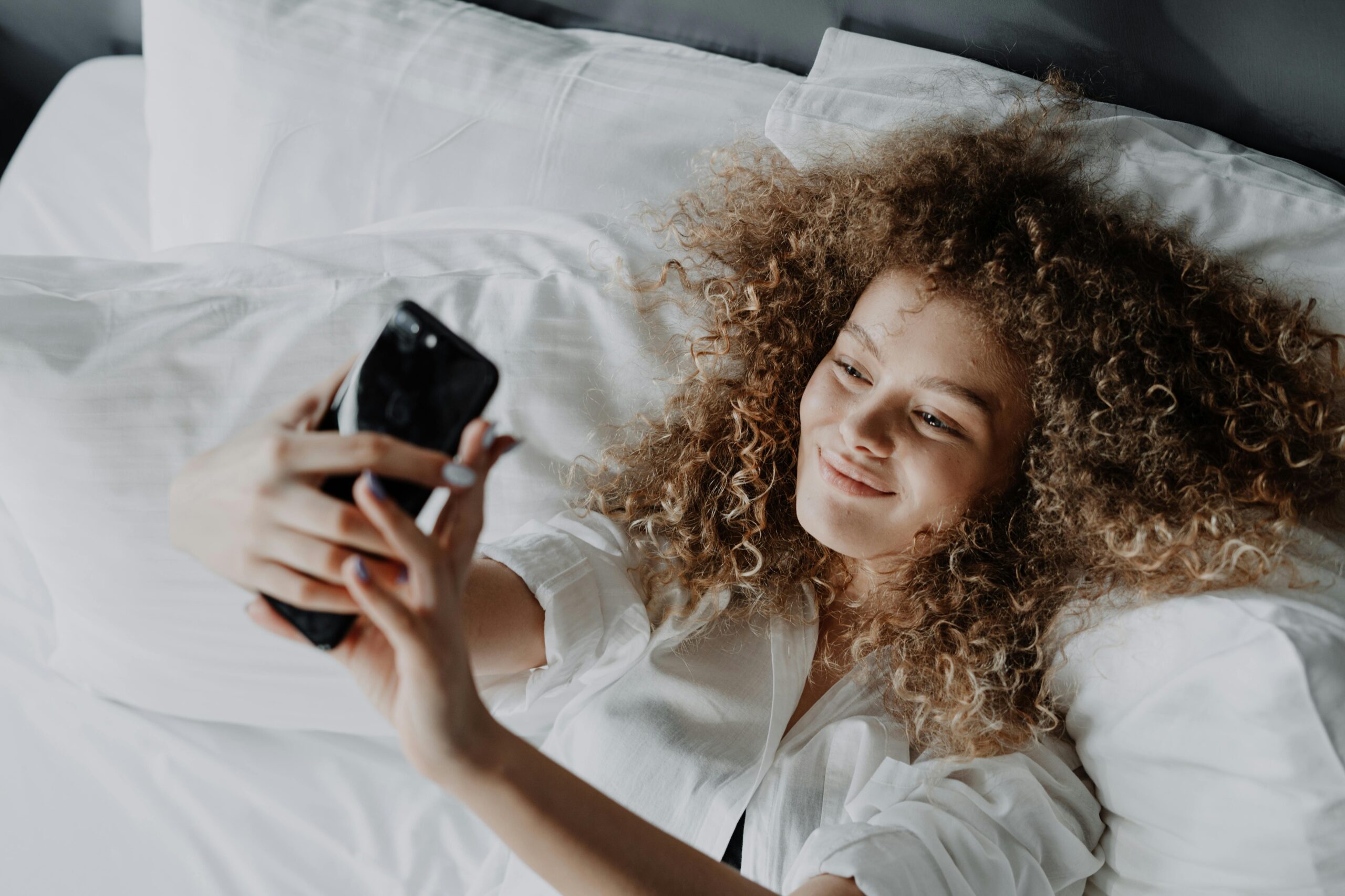 A woman in a white button up shirt lying on a bed with white sheets