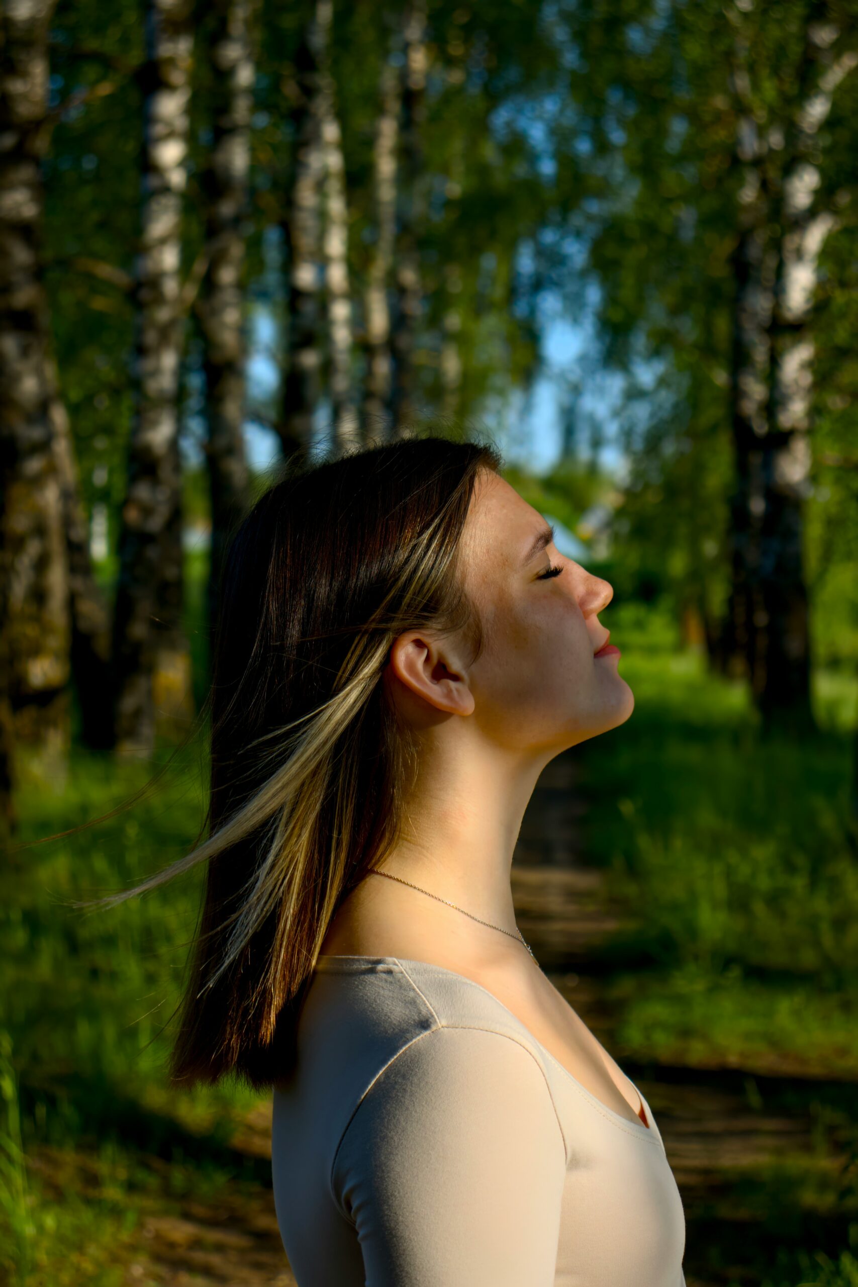 A woman with blonde hair wearing a tan top with her eyes closed in a forest