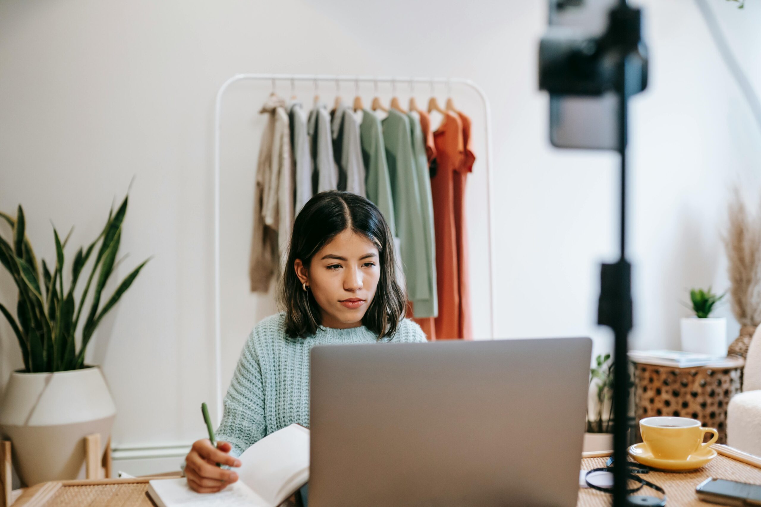 A woman with short black hair and a wearing blue sweater writing in a notebook with her laptop open. In the background is a clothing rock with grey, green, and orange long-sleeve shirts and dresses. 