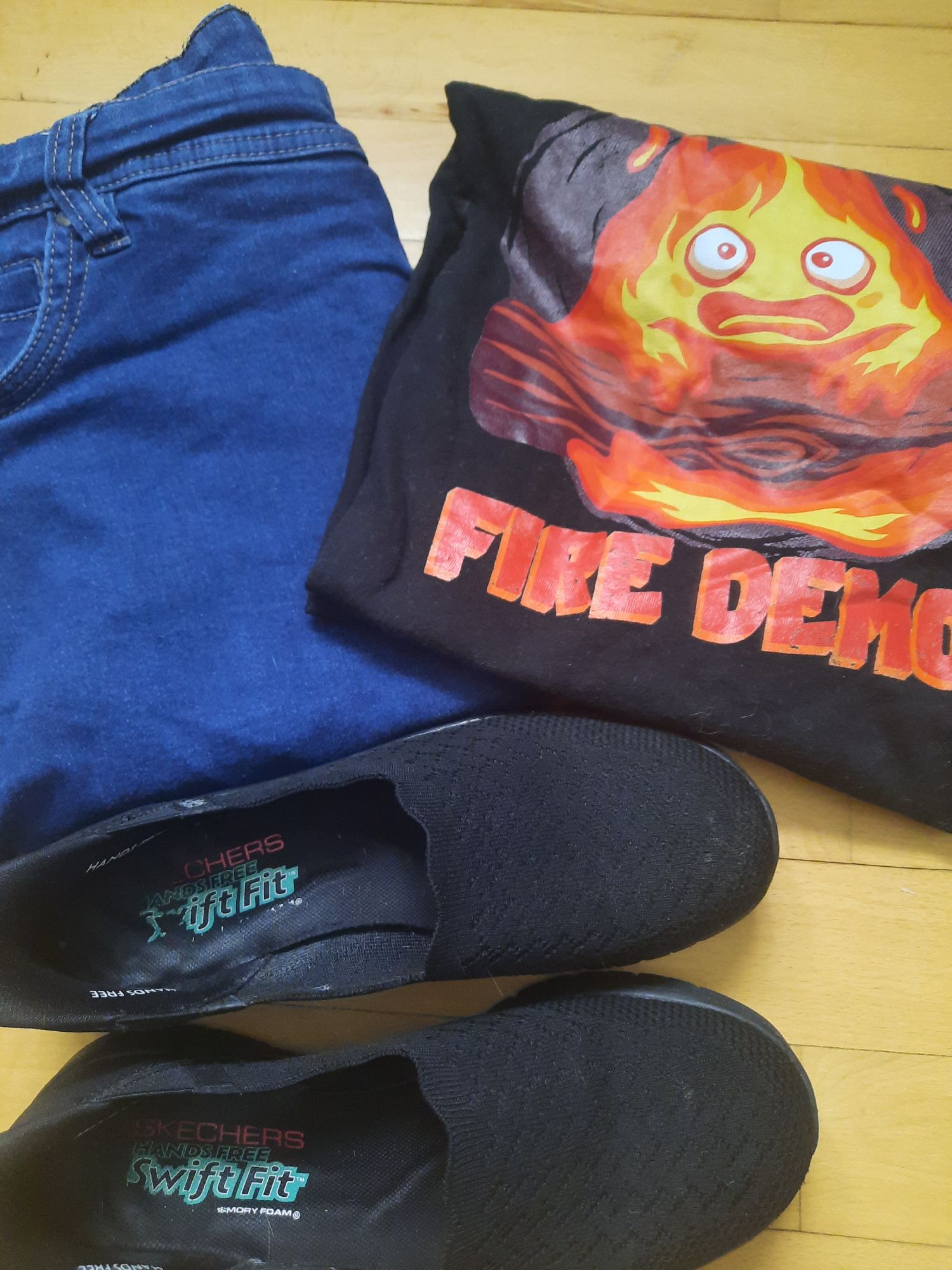 My blue jeans folded next to the black Calcifer print t-shirt with the saying "Emotional Support Fire Demon" and my black Sketchers slip-ins a