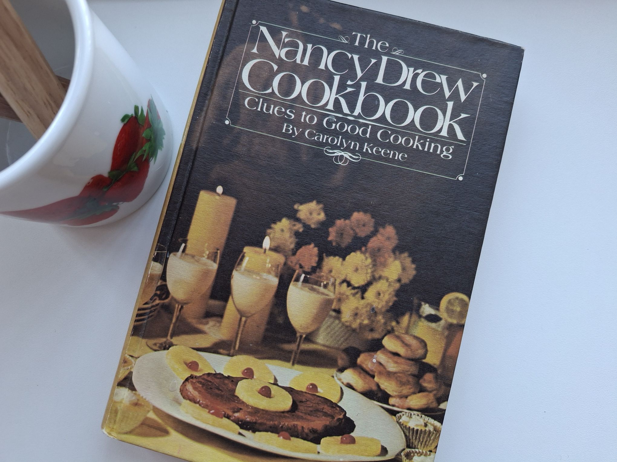 The Nancy Drew Cookbook next to a white container with strawberries on it
