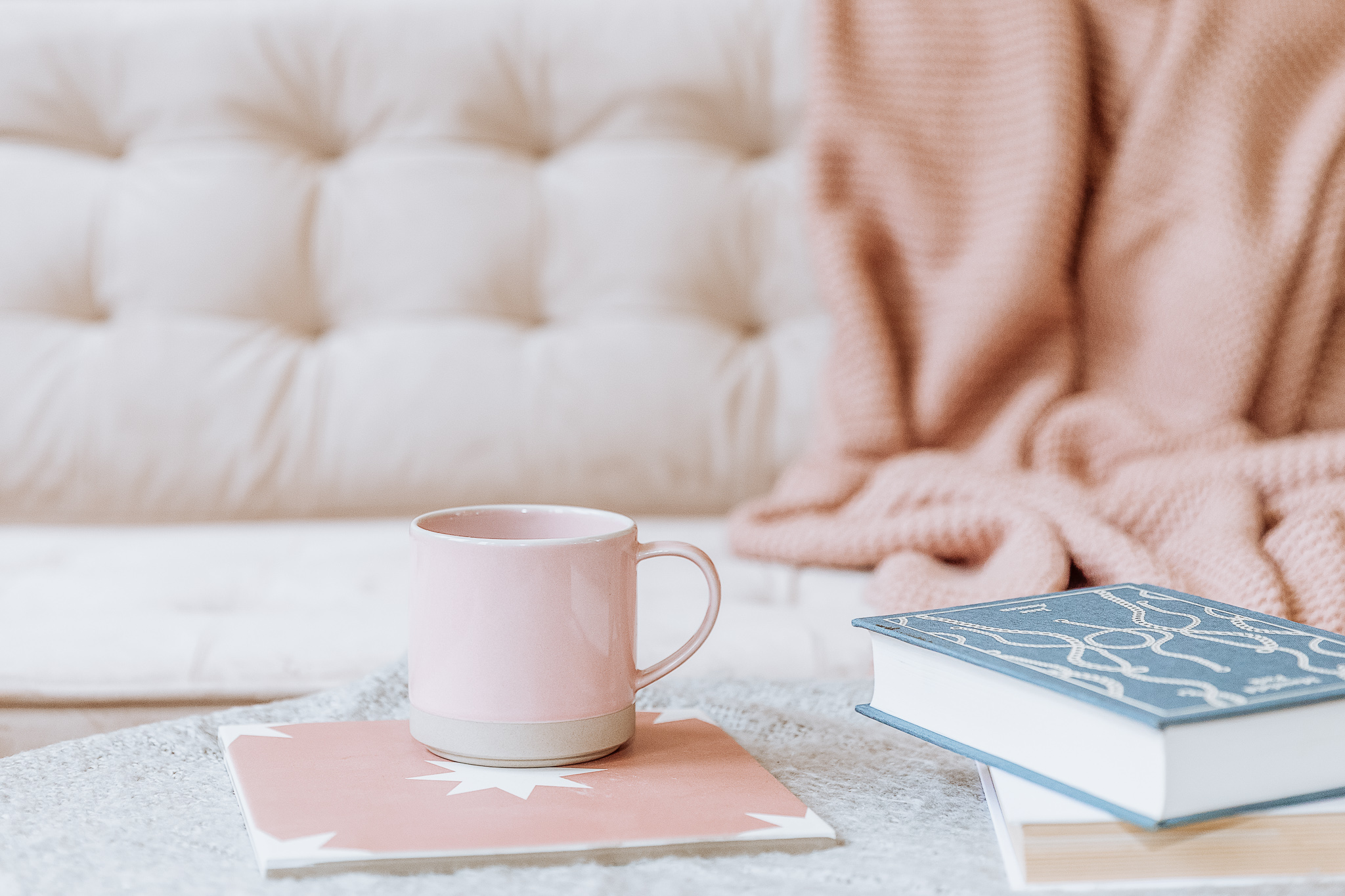 A stack of books and a pink mug on a coffee table in front of a couch with a blanket