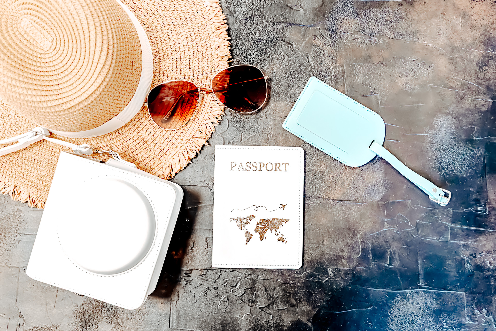 A flat lay of a straw sunhat, sunglasses, white bag, passport cover, and blue luggage tag.