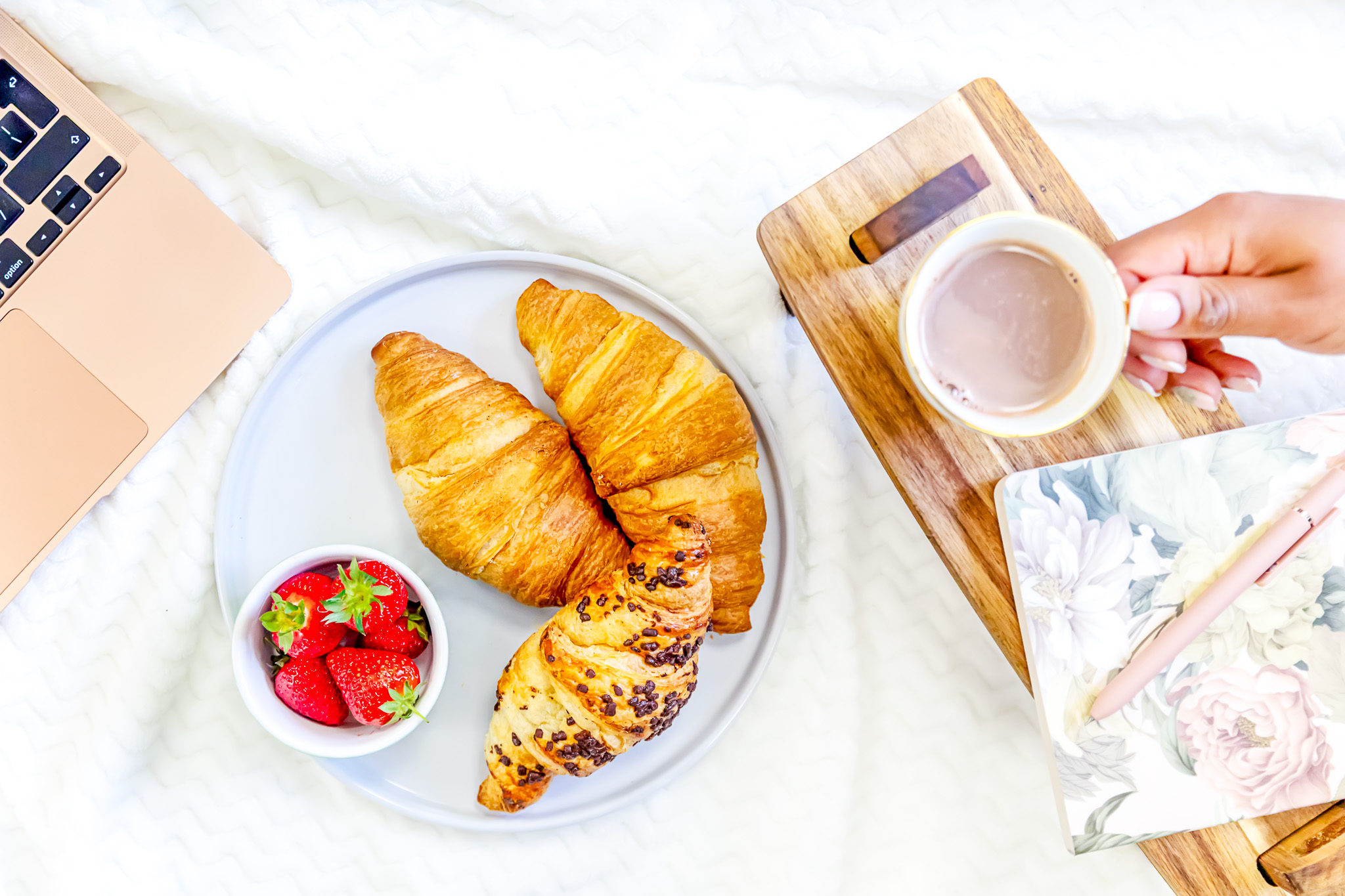 A plate of croissants and strawberries with a mug of hot chocolate next to a laptop and notebook with a pen