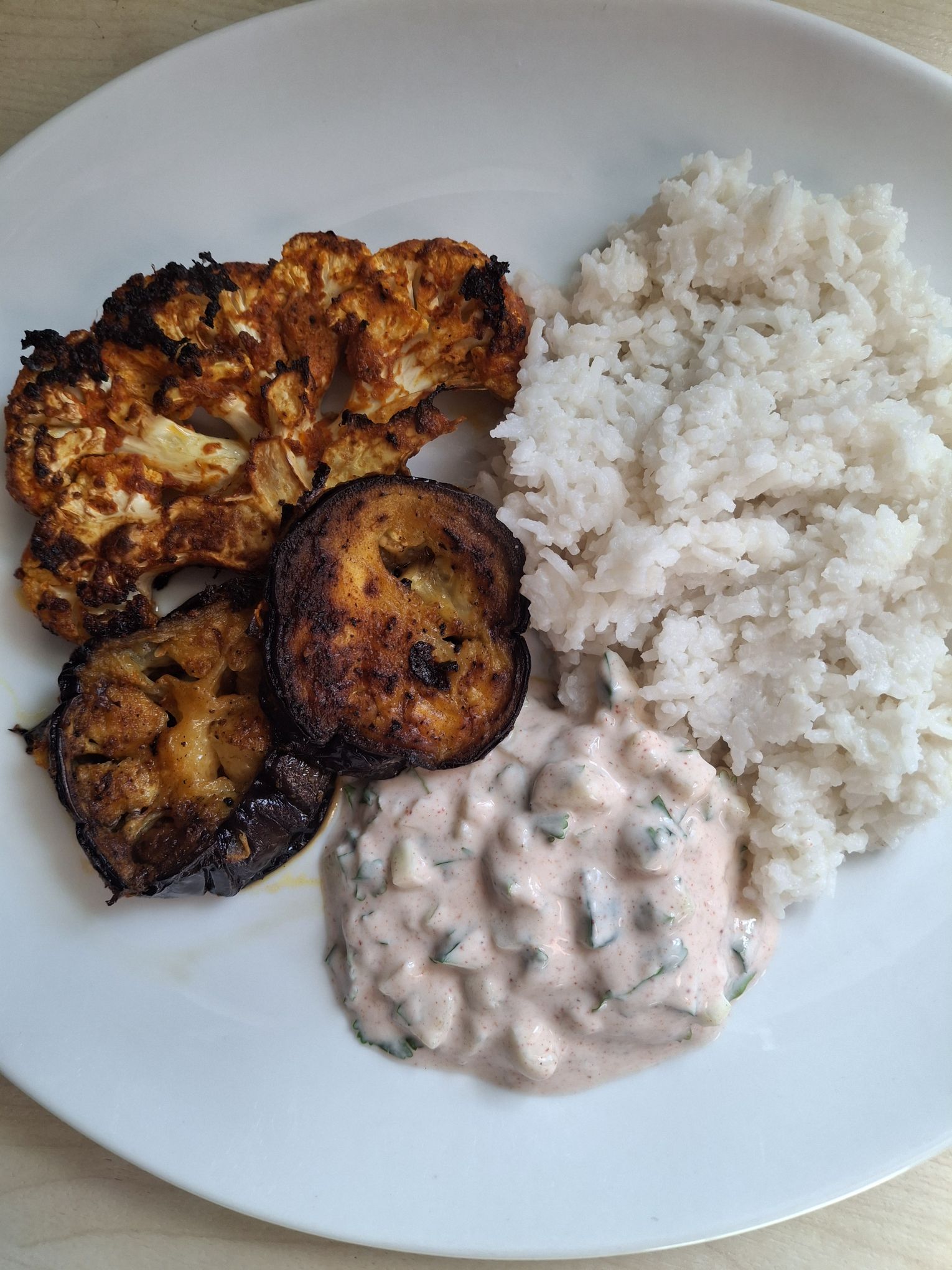 The tandoori cauliflower and eggplant steaks on a plate with a cucumber raita and rice from The Official Bridgerton Cookbook